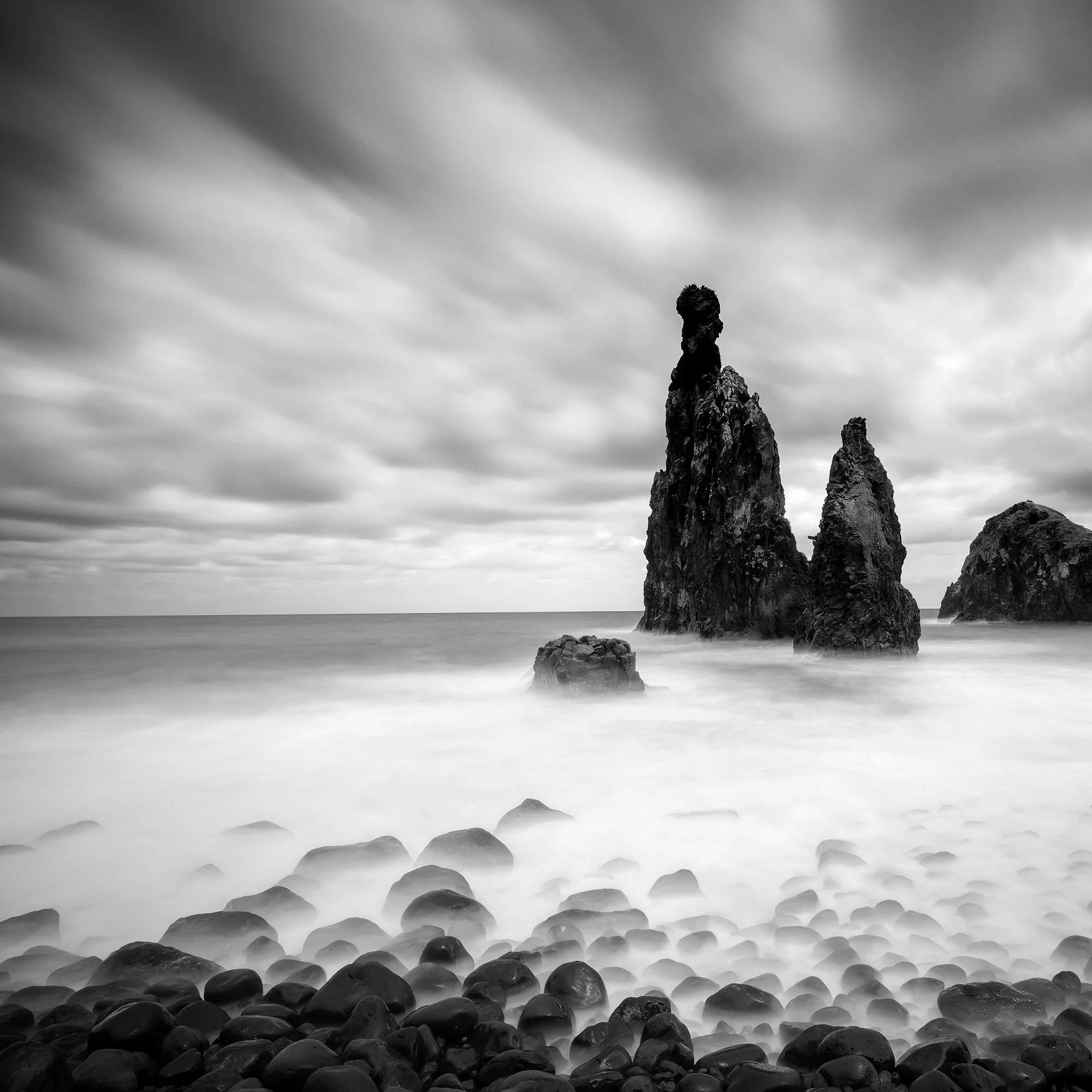 © 2021 Gerald Berghammer - Black and white seascape photography. Rocky seashore with three prominent sea stacks rising from the water, cloudy sky overhead. Print detail 1
