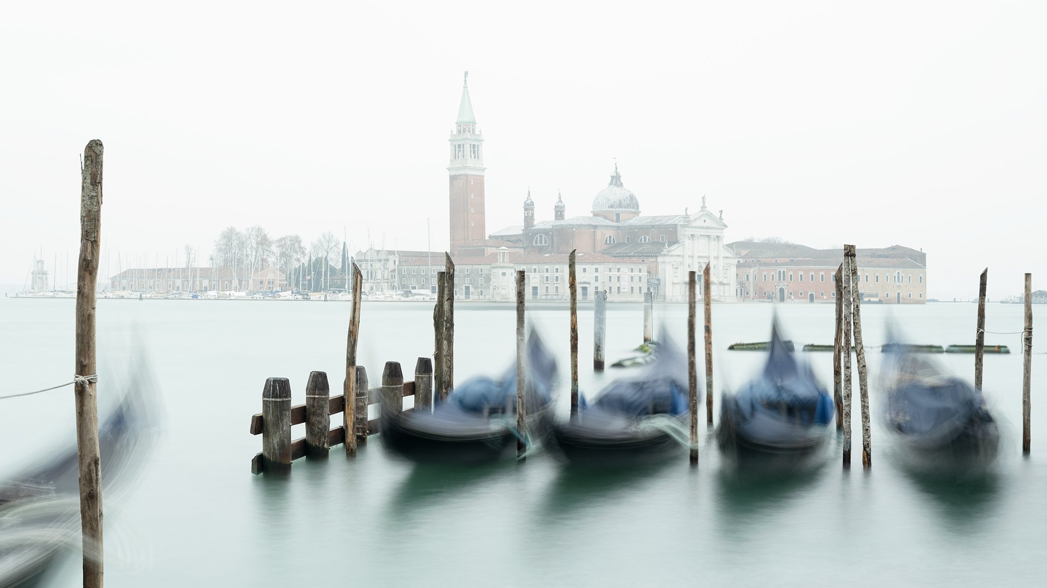 Gondolas moving beside wooden mooring posts with San Giorgio Maggiore in the misty background, Venice. Photo by Gerald Berghammer.