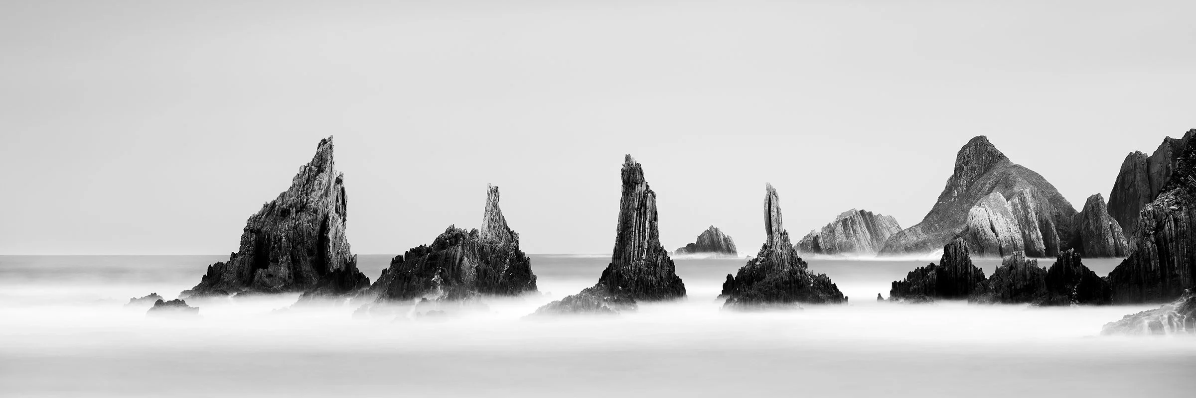 Monochrome coastal landscape featuring dramatic rock formations emerging through soft sea mist.
