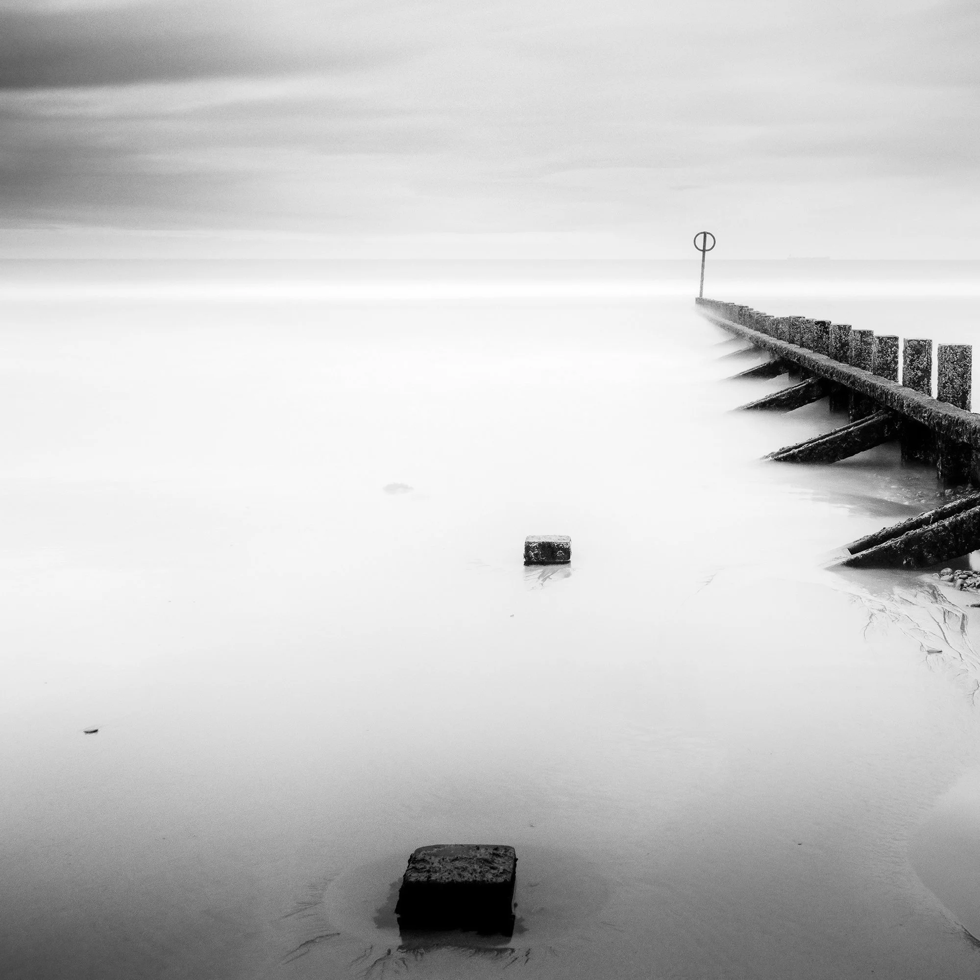 © 2012 Gerald Berghammer - Black and white landscape photography. A wooden wave breaker extending into a calm body of water, with a circular marker at the end. Print detail 1