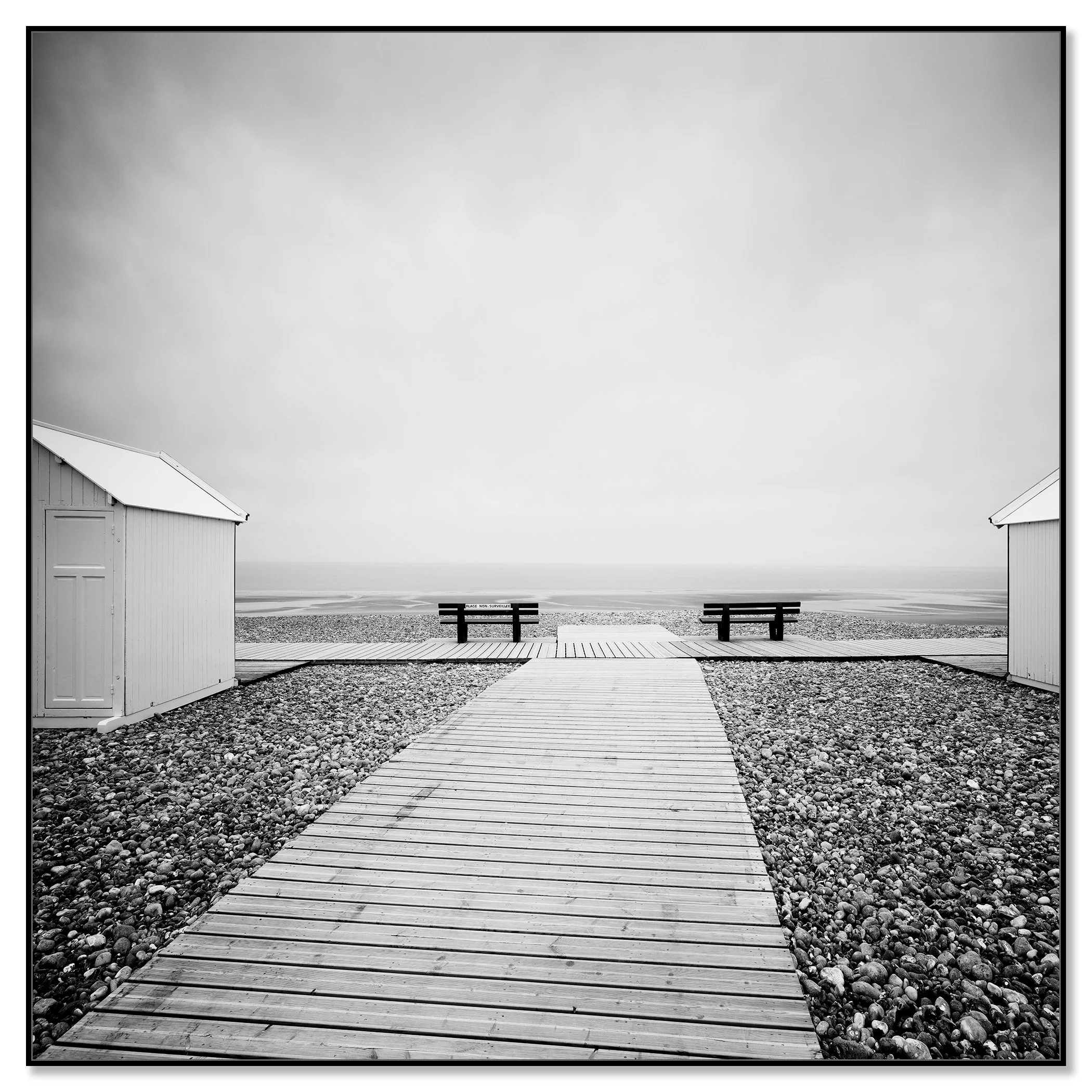Black and white photo of a wooden boardwalk leading across a pebble beach to benches facing the sea – framed ArtBox black