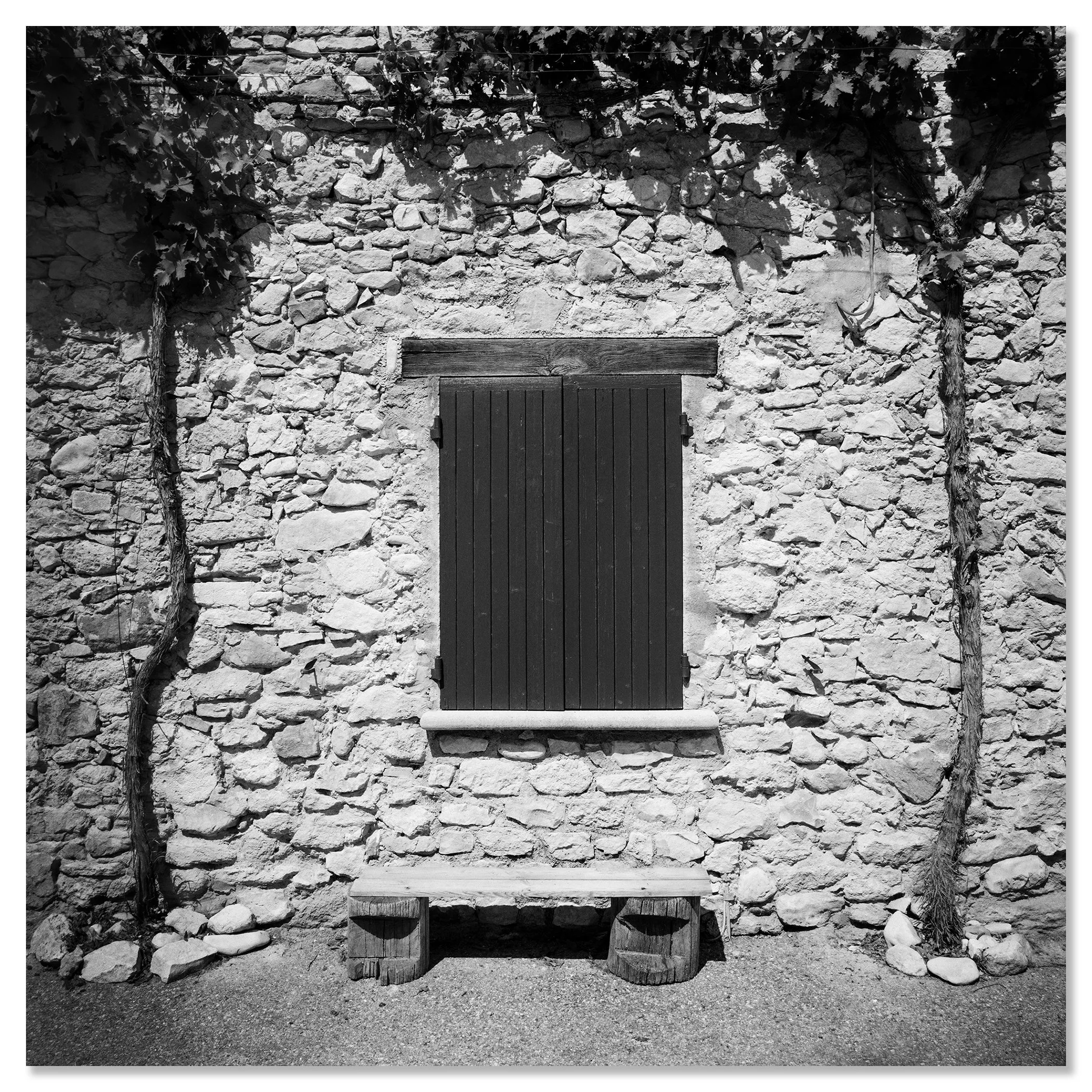 Gerald Berghammer - Black & white photography. Stone wall with a closed wooden window shutter, flanked by climbing vines on both sides, and a small bench. Chromaluxe frameless