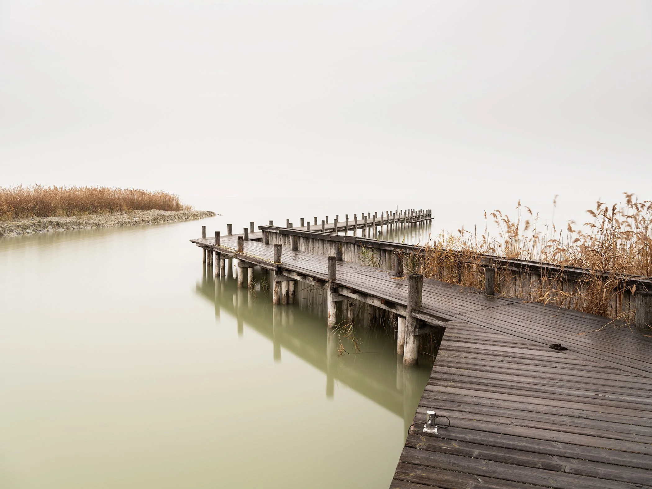 Minimalist colour photograph of a weathered wooden jetty extending into misty Lake Neusiedl, Austria, by Gerald Berghammer.