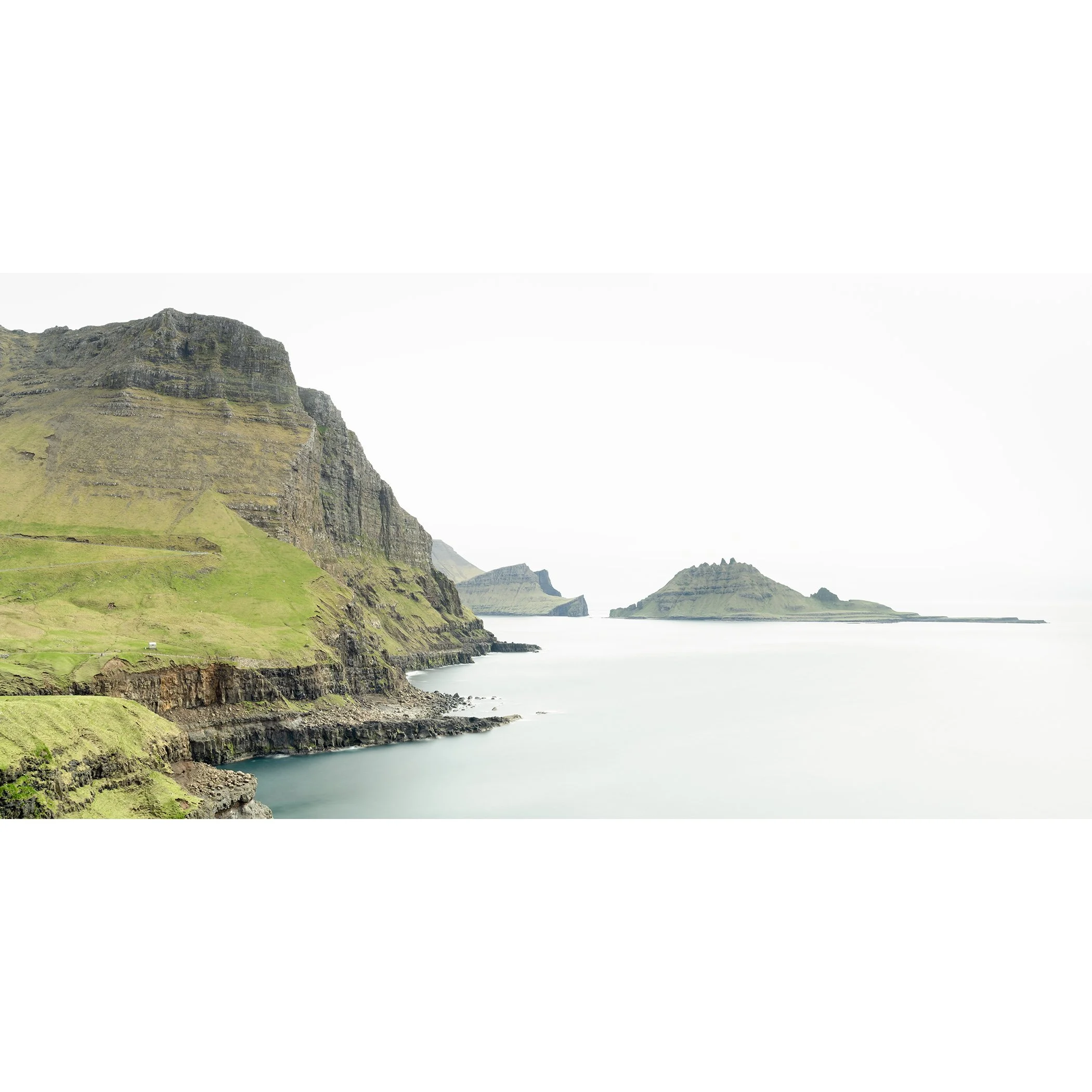Gerald Berghammer - Color Photography. Cliffs and green hills along a body of water, with distant islands or landmasses in the background under a cloudy sky.