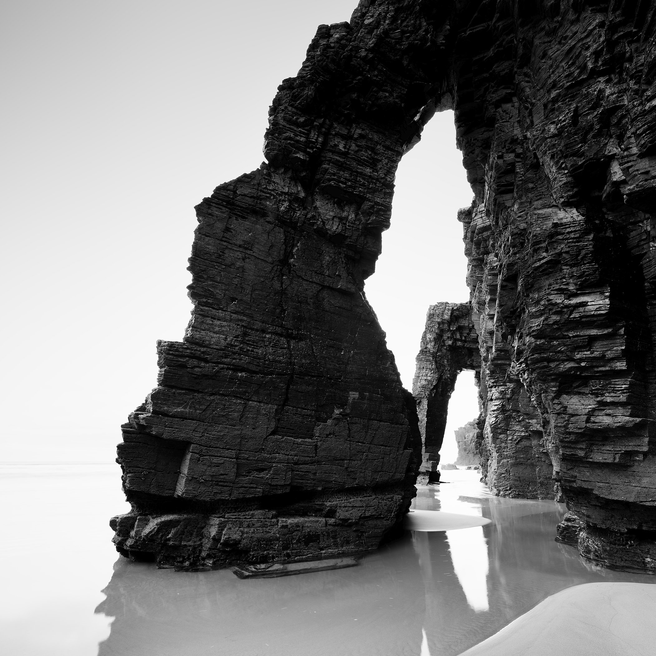 Natural stone arches and dark coastal cliffs on a calm beach in a minimalist black and white scene