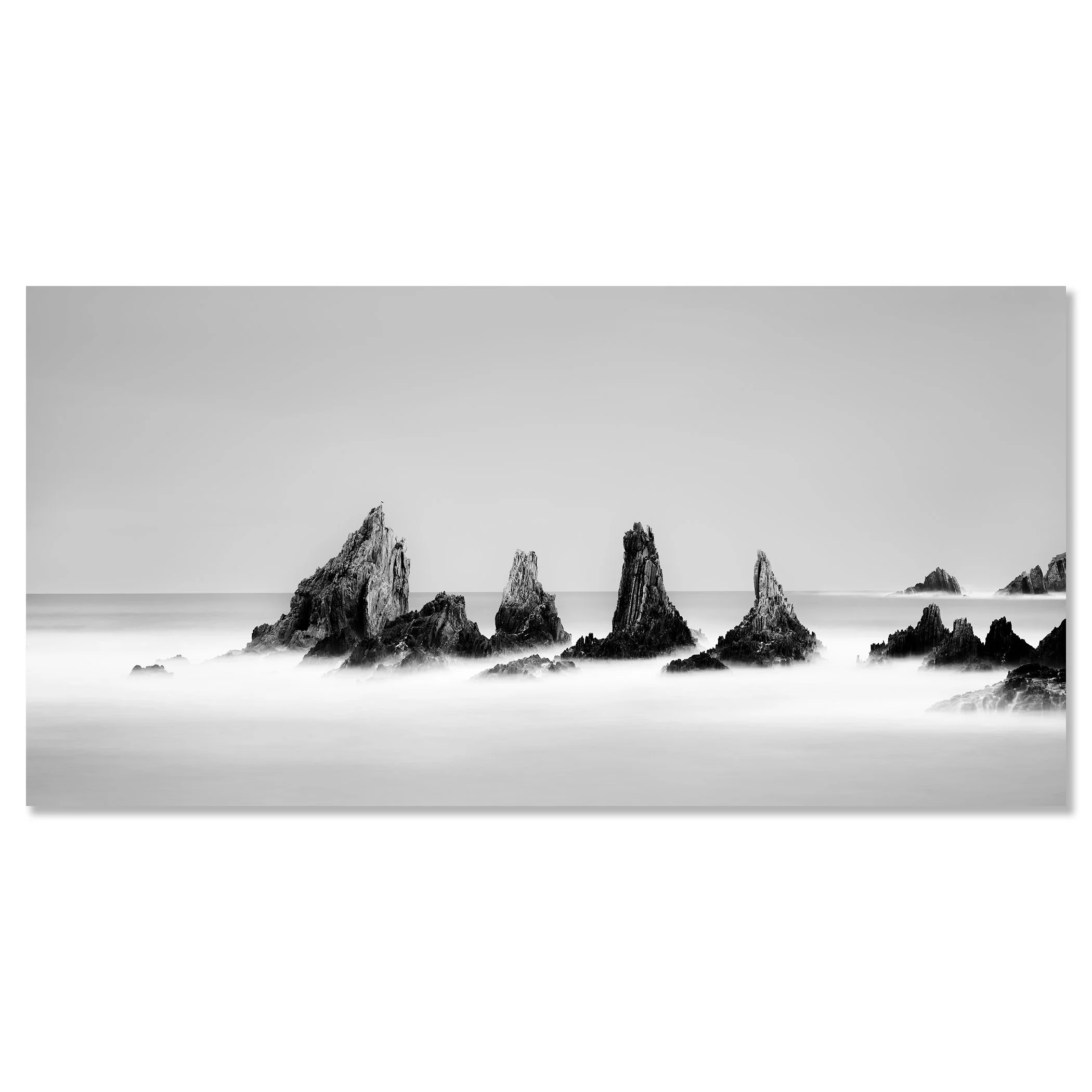 Black-and-white long-exposure photograph of jagged rocks in calm ocean water, misty waves and clear sky – dibond frameless