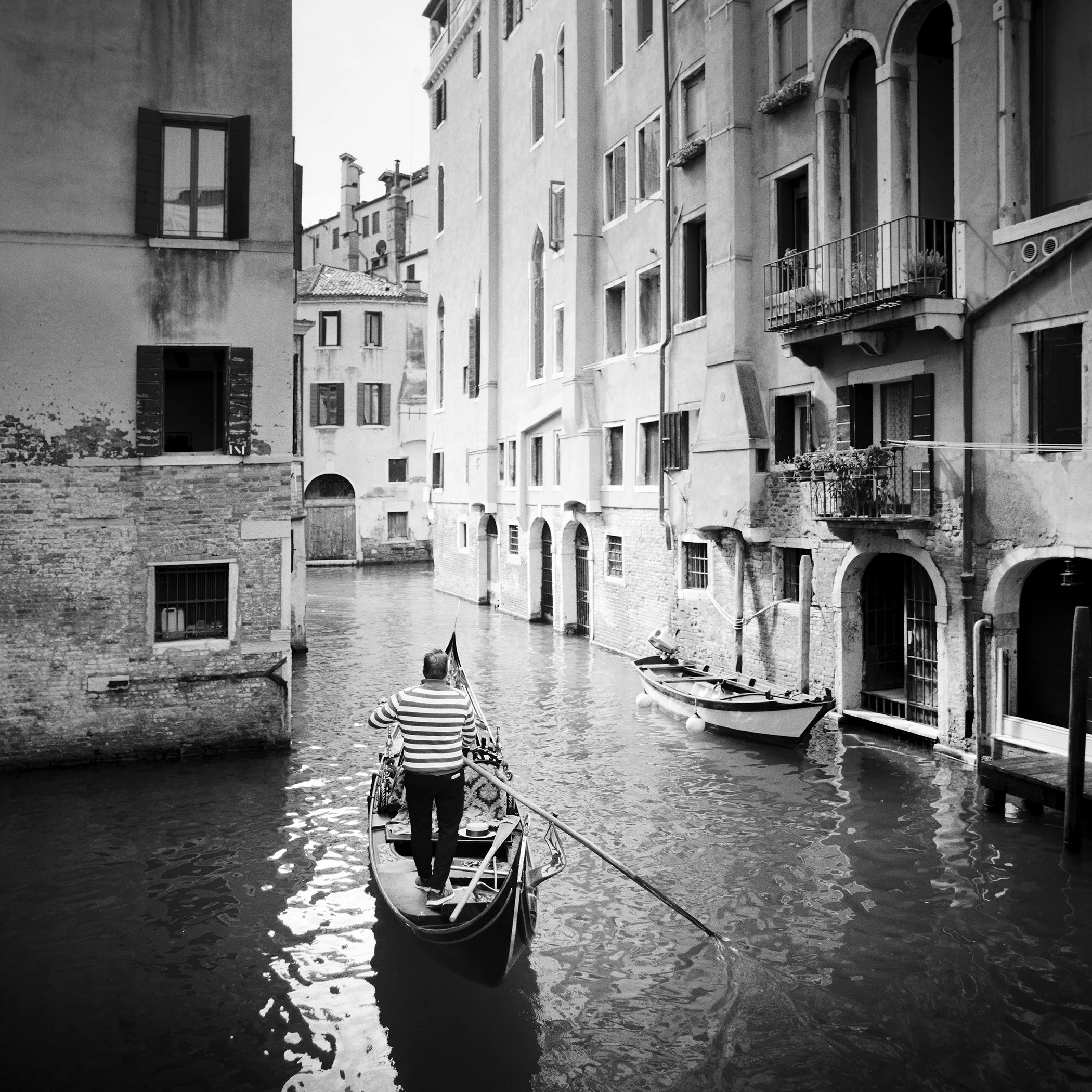Monochrome photo of a gondolier steering through a narrow Venetian canal between historic buildings in Venice