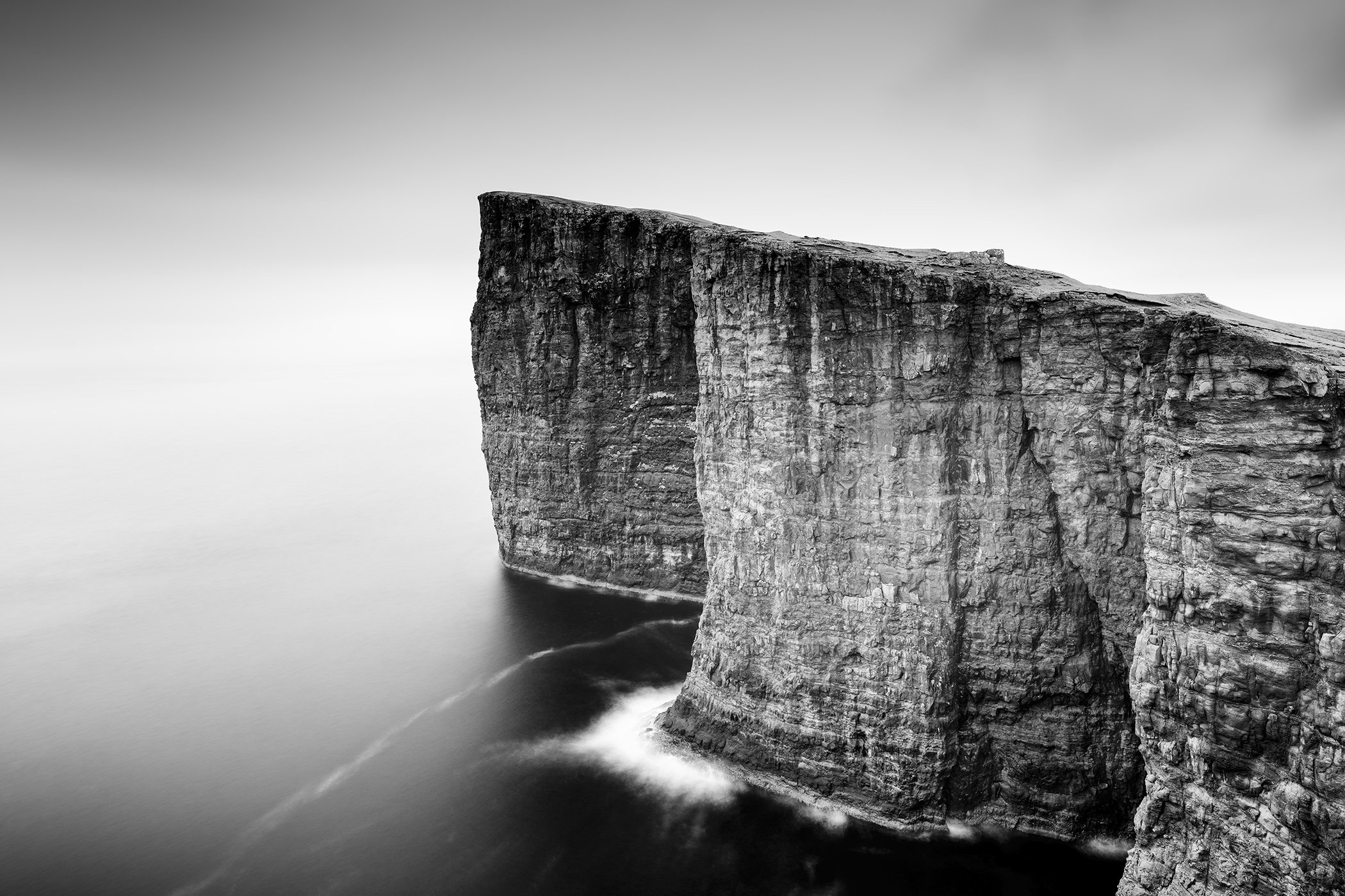 © 2024 Gerald Berghammer. Black-and-white photograph of a massive coastal cliff rising vertically from calm water, with long-exposure waves creating a soft, mist-like effect at the base under an overcast sky.
