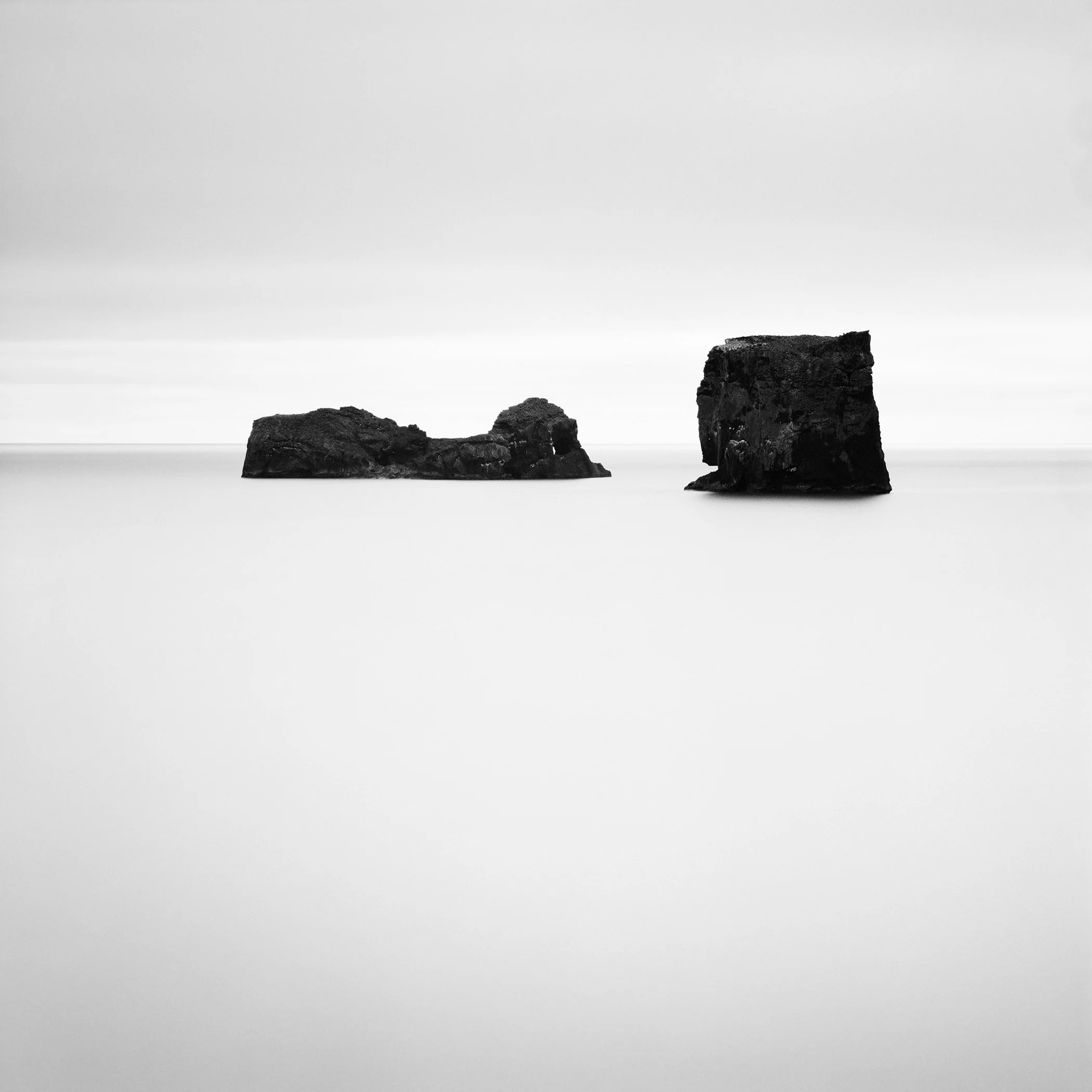 Two dark rock formations rising from a calm, mist-like sea in a minimalist black-and-white seascape in Iceland. Photo by Gerald Berghammer.