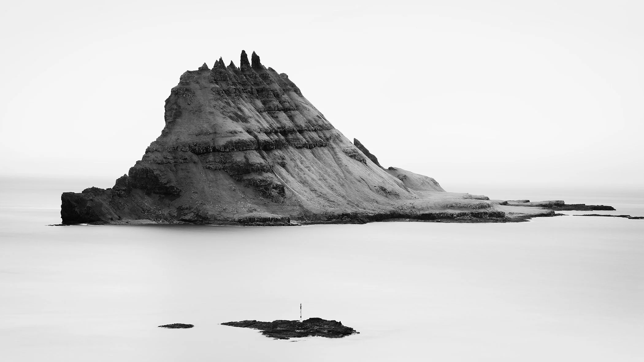 Black-and-white minimalist photo of a solitary jagged sea stack rising from calm misty ocean water, with a soft fading horizon.