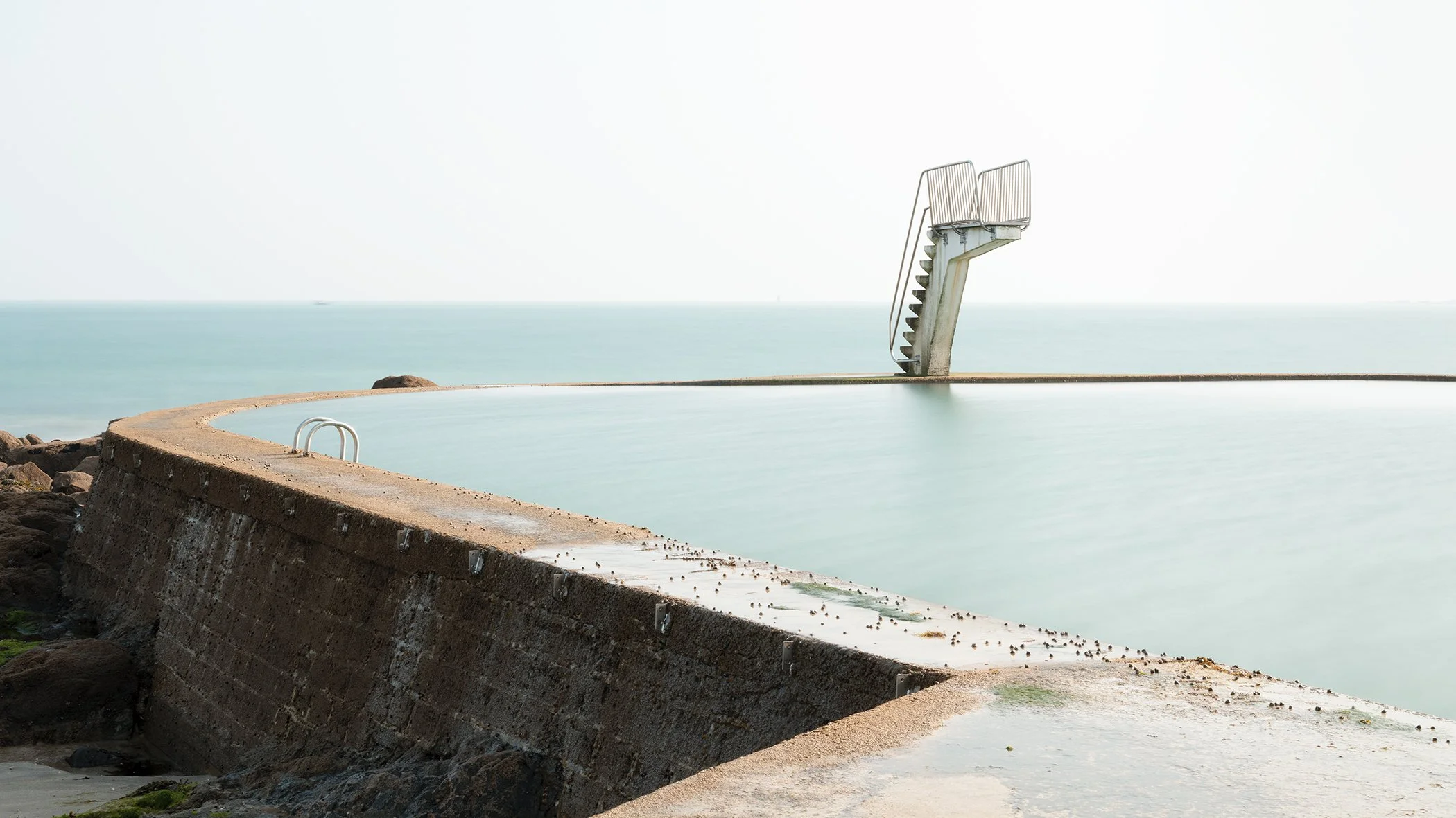 Minimalist colour photograph of a seawater swimming pool with diving platform on the French coast, photo by Gerald Berghammer.