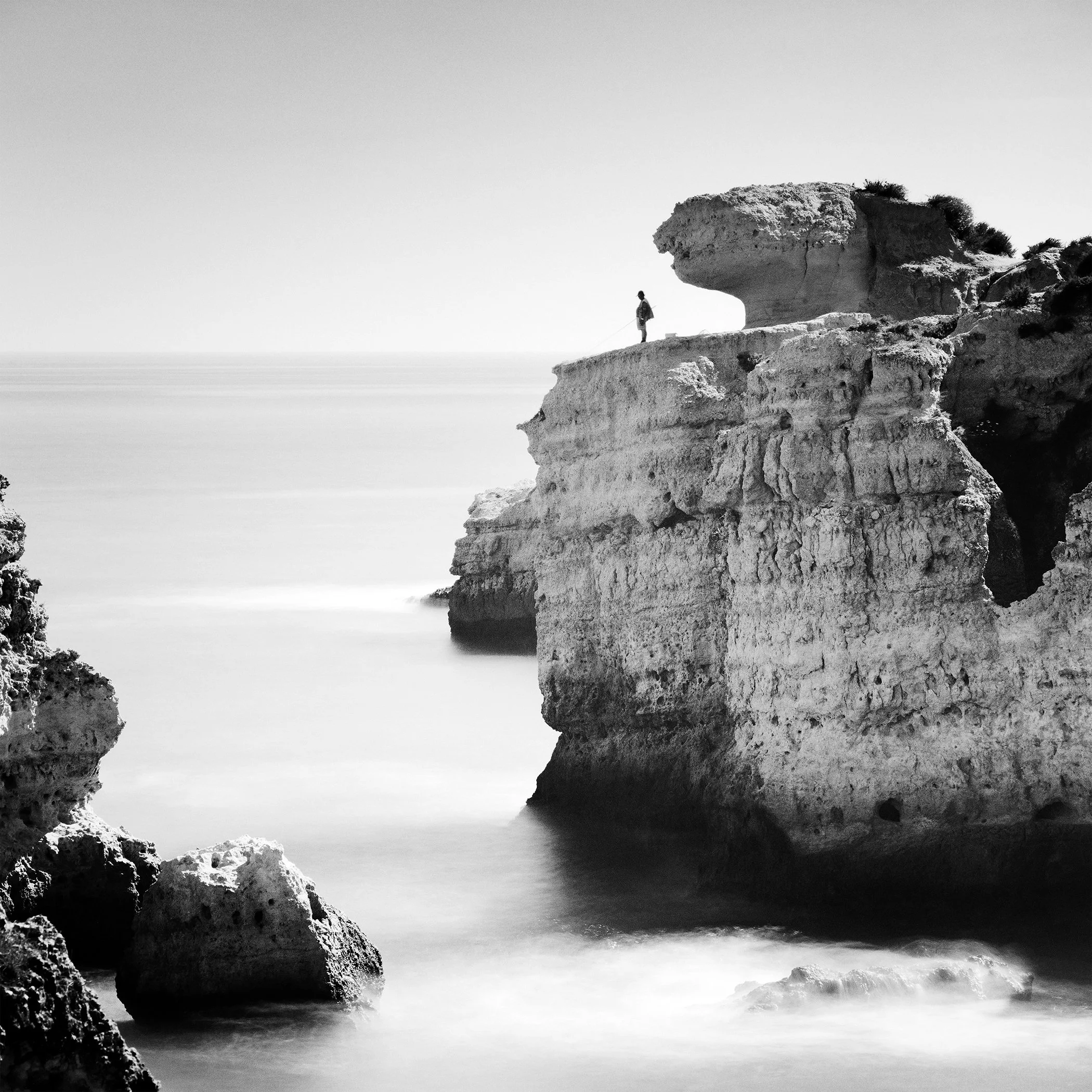 Black-and-white long-exposure photograph of a lone fisherman standing on layered coastal cliffs above calm water in Portugal. Photo by Gerald Berghammer.
