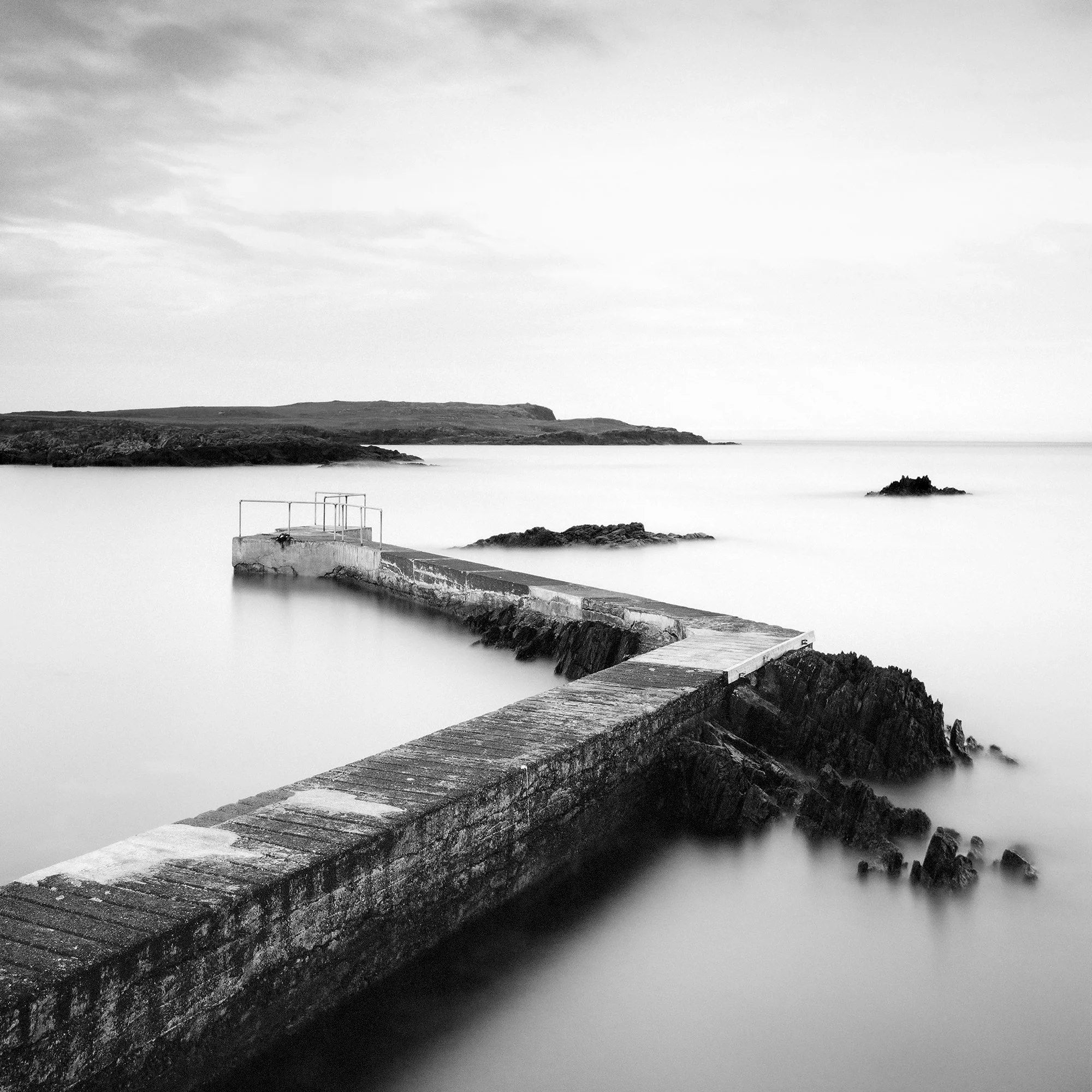 © 2014 Gerald Berghammer - Black and white seascape photography. A Concrete pier extends into calm seawater with rocky edges, under a cloudy sky. Print detail 1