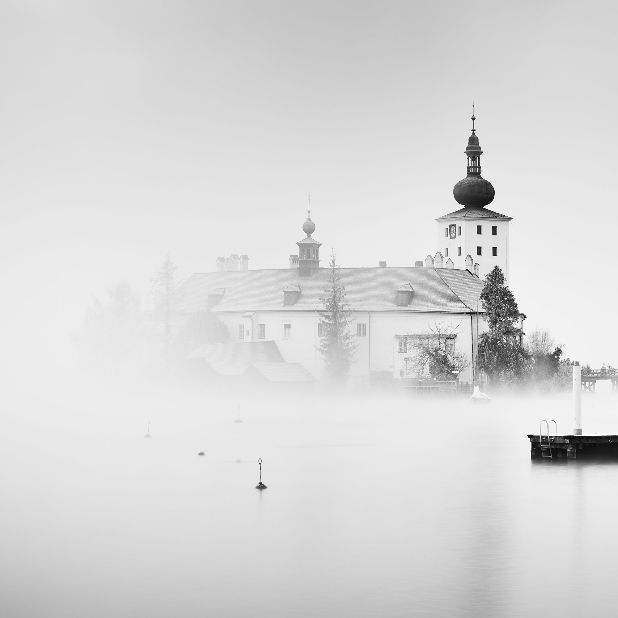 © 2022 Gerald Berghammer - Black and white cityscape photography. Schloss Ort - The historic church surrounded by mist over a lake with docks in the foreground. Print detail 1