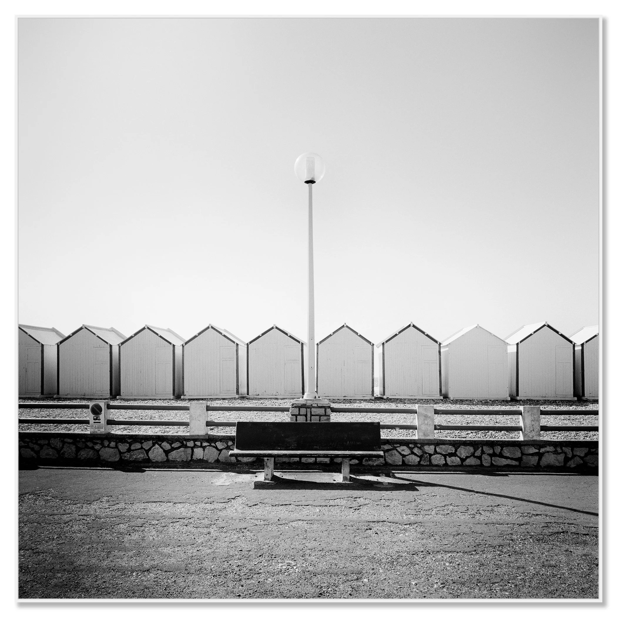 Empty bench on the promenade facing beach huts, centred streetlamp, minimalist black-and-white coastal photograph, ChromaLuxe frame white