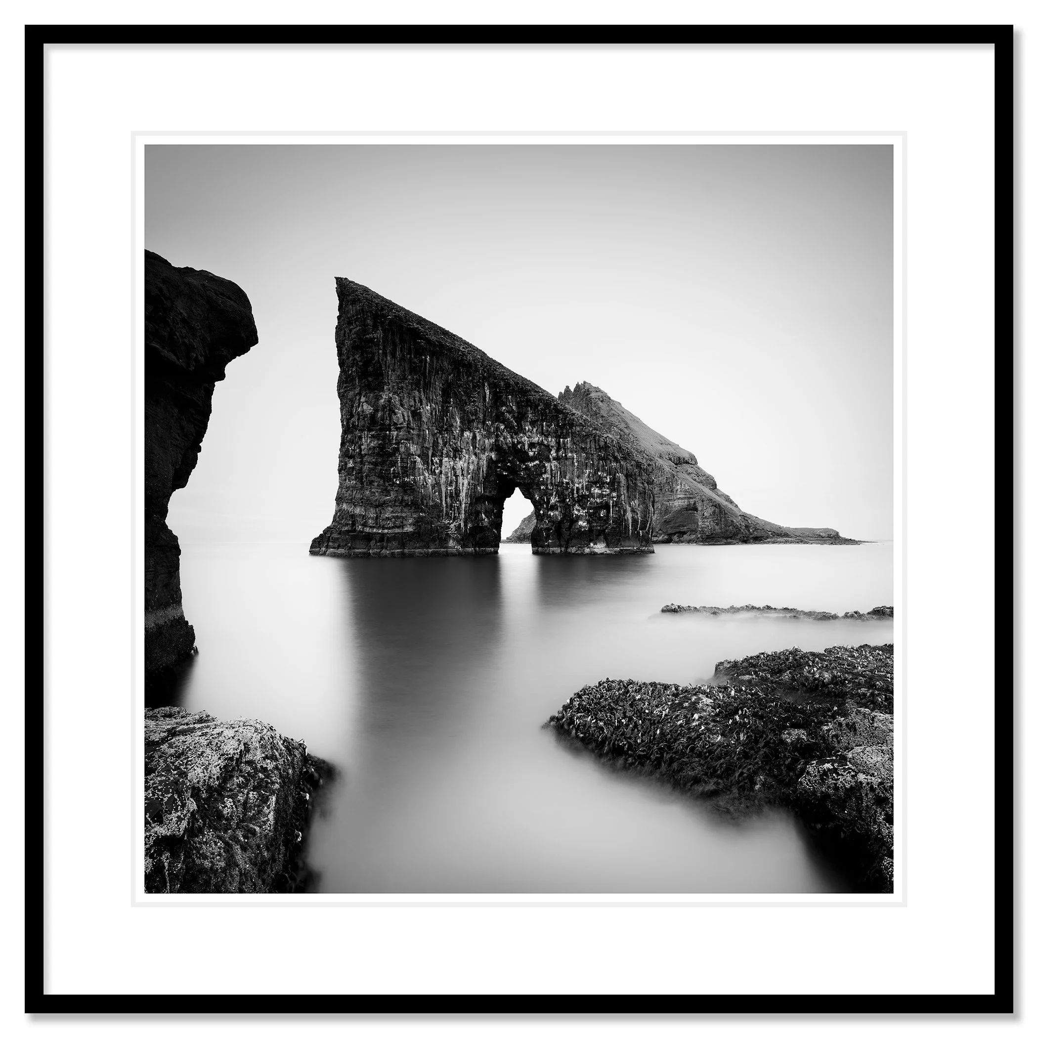 Black and white coastal landscape with a striking sea stack arch and soft long-exposure water – classic framed black