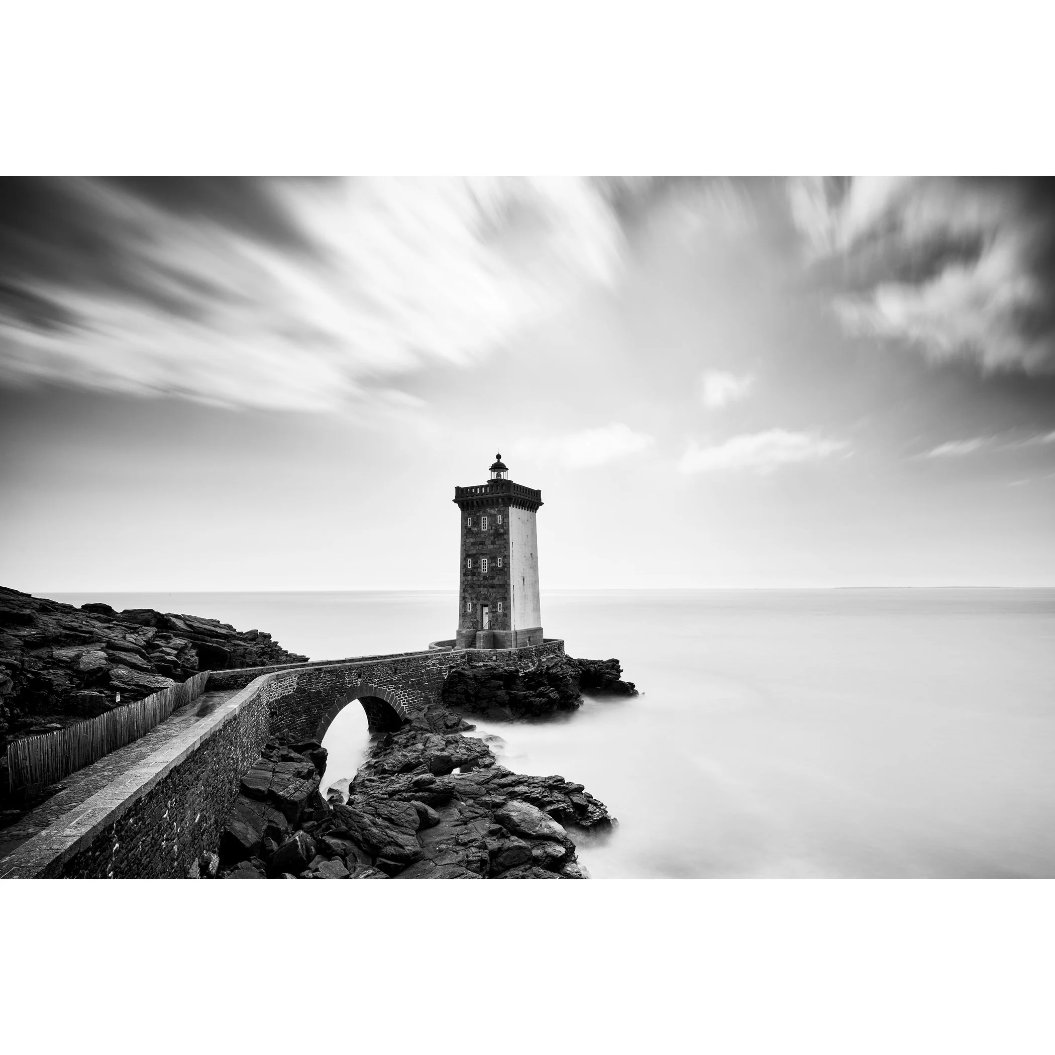 Black-and-white coastal lighthouse with stone bridge across rocks and overcast sky