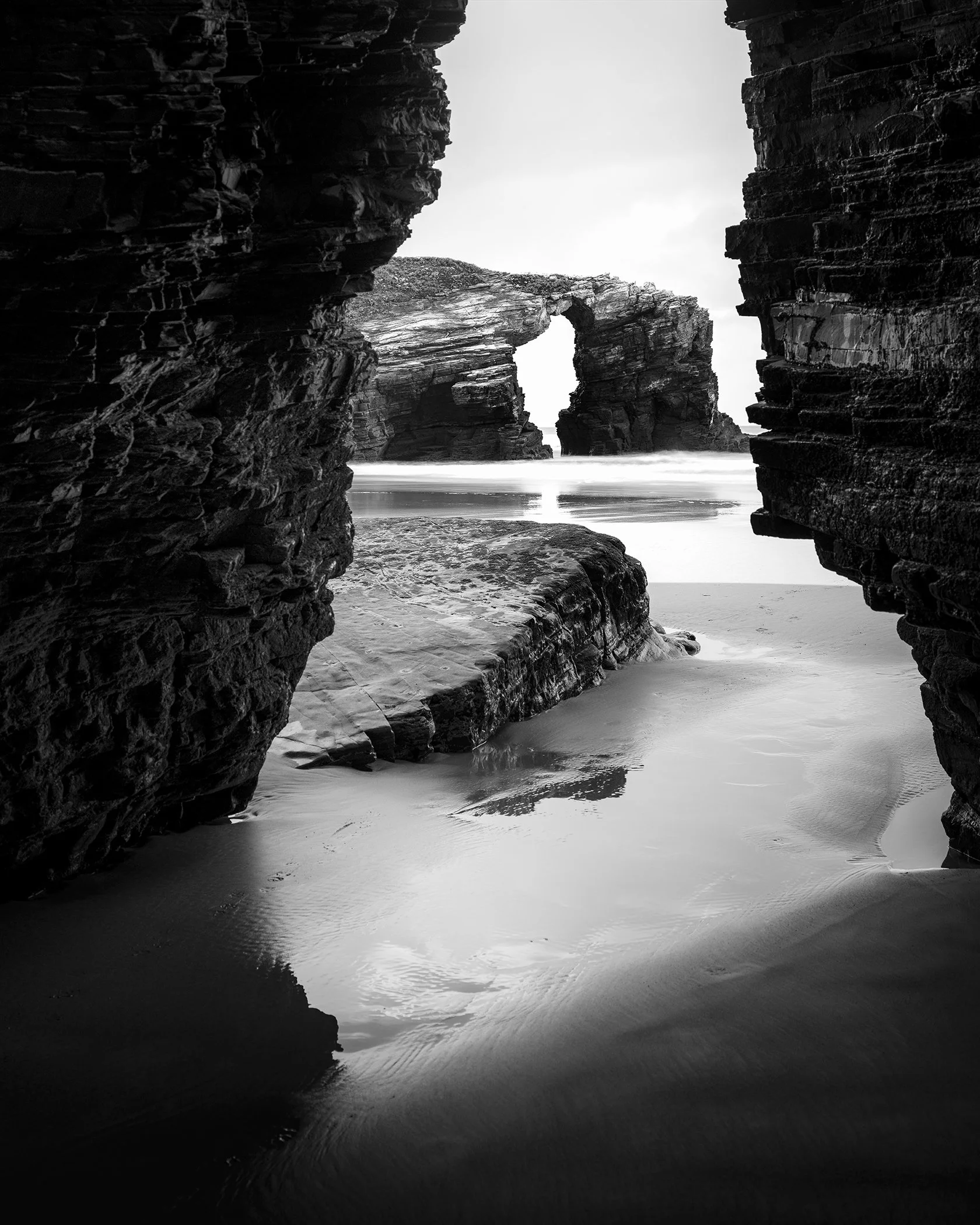 Black and white landscape photograph of a rocky beach in Spain with natural rock arches, framed through a narrow gap between large coastal rocks.