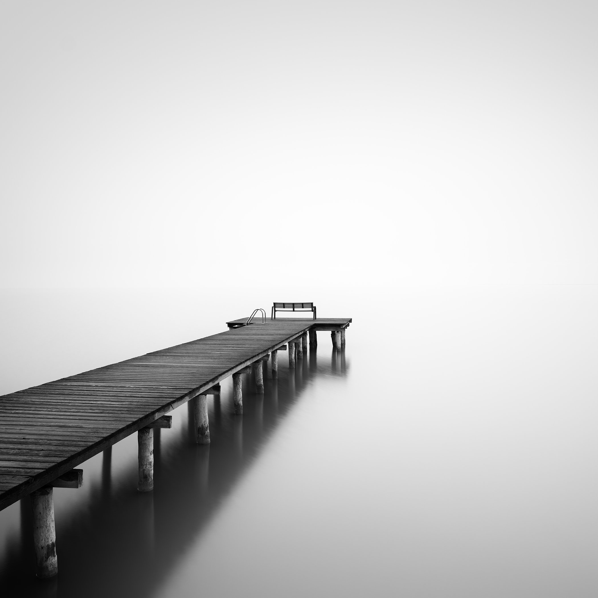 © 2025 Gerald Berghammer - Minimalist black and white photograph of a wooden pier extending into calm, misty water, creating a serene and contemplative atmosphere.