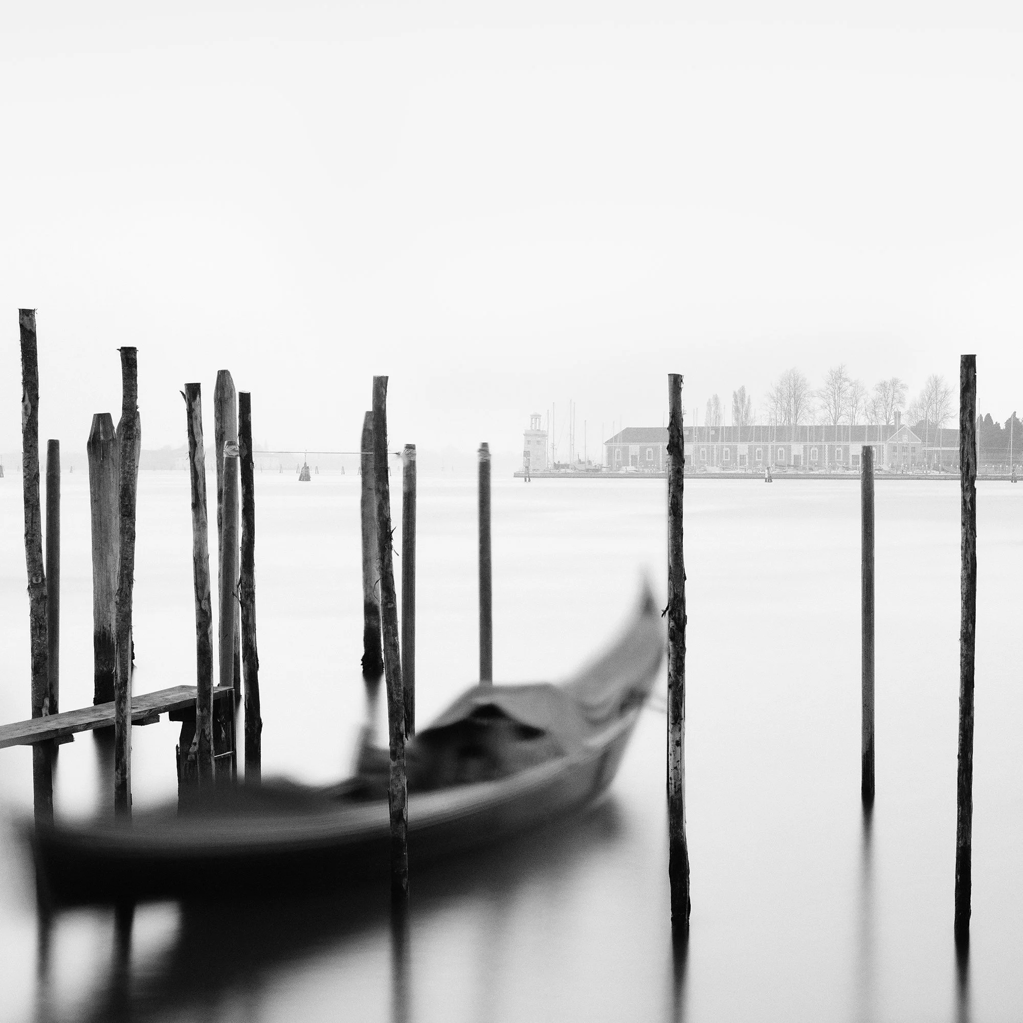 Gerald Berghammer - Black and white cityscape photography. Gondolas moored along wooden posts on a canal with view of a city skyline and a tall bell tower. Print detail 1