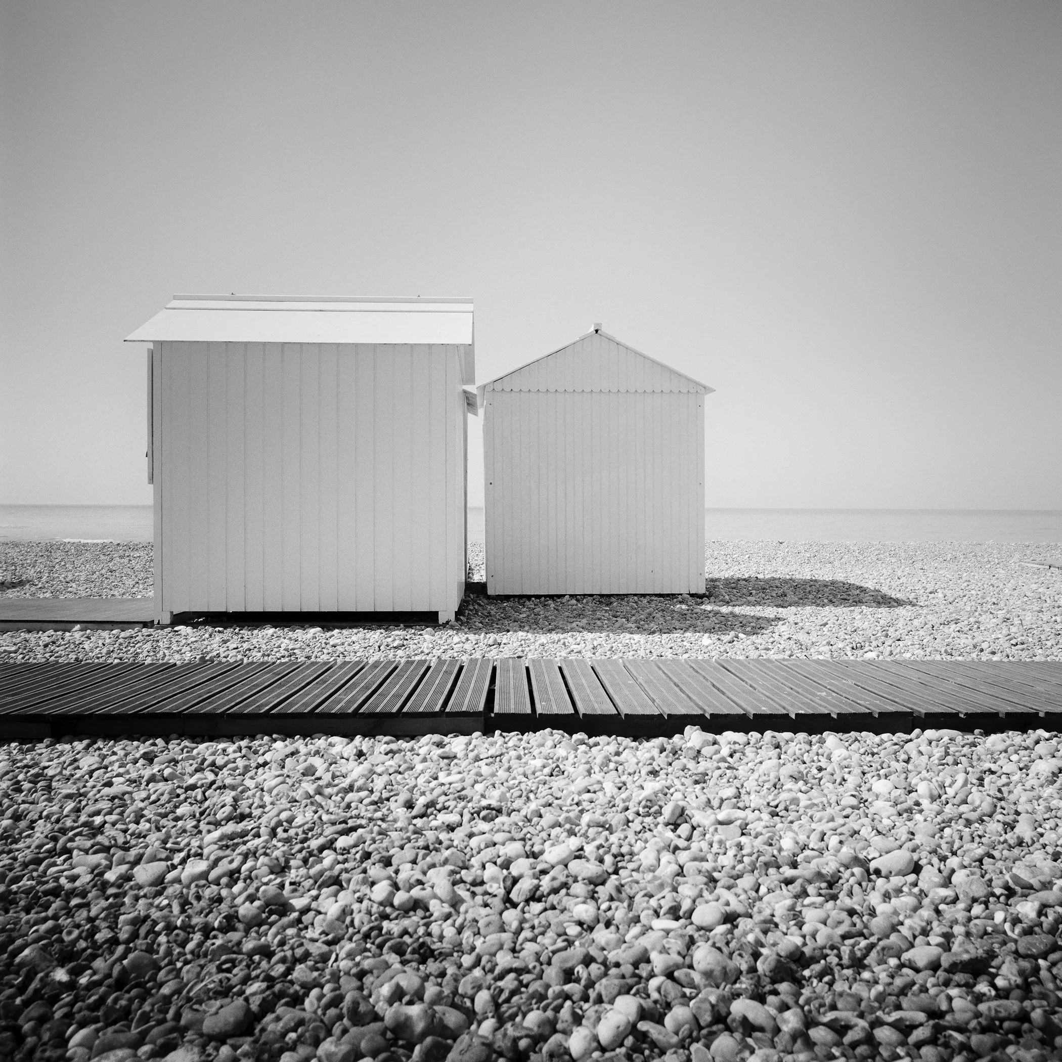 Fine art black and white beach photograph by Gerald Berghammer showing two white cabins on a quiet pebble coastline