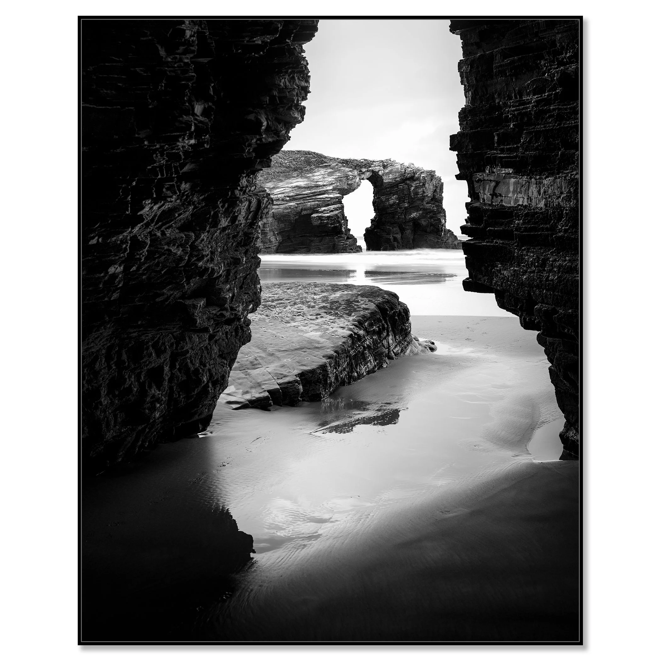Monochrome seascape photo of Catedrais Beach, Spain, showing a natural rock arch and ocean waves viewed through a narrow opening between large rocks – framed ArtBox black