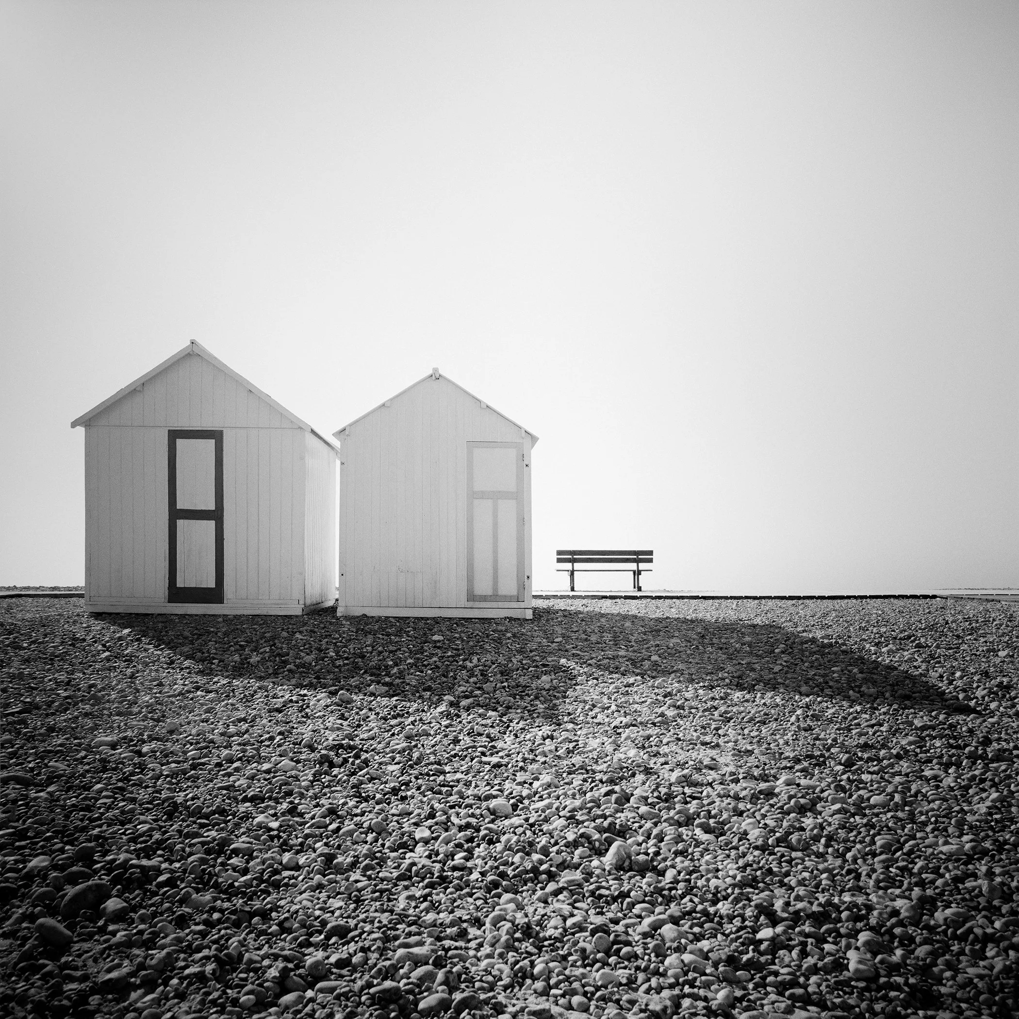 Two white beach huts and a solitary bench on a misty pebble beach in France. Black-and-white photo by Gerald Berghammer.
