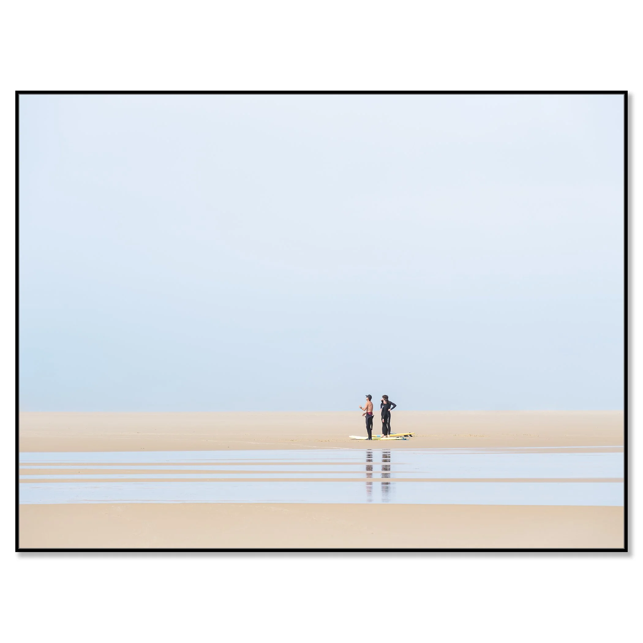 Gerald Berghammer - Color landscape Photography. People stand on a sandy beach near the ocean, wearing wetsuits and surfboards against a pale blue sky. Chromaluxe framed black