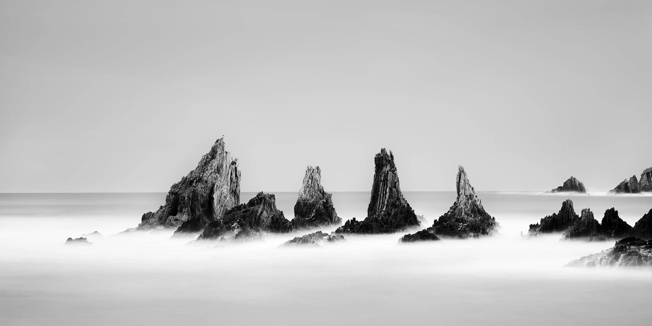 Long-exposure black-and-white ocean scene with jagged rocks, smooth misty water and clear sky