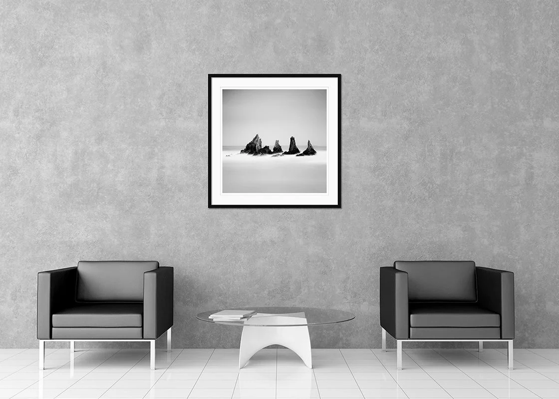 Minimalist living room with two black armchairs, a glass coffee table, and a framed black-and-white landscape photograph by Gerald Berghammer on a gray wall.
