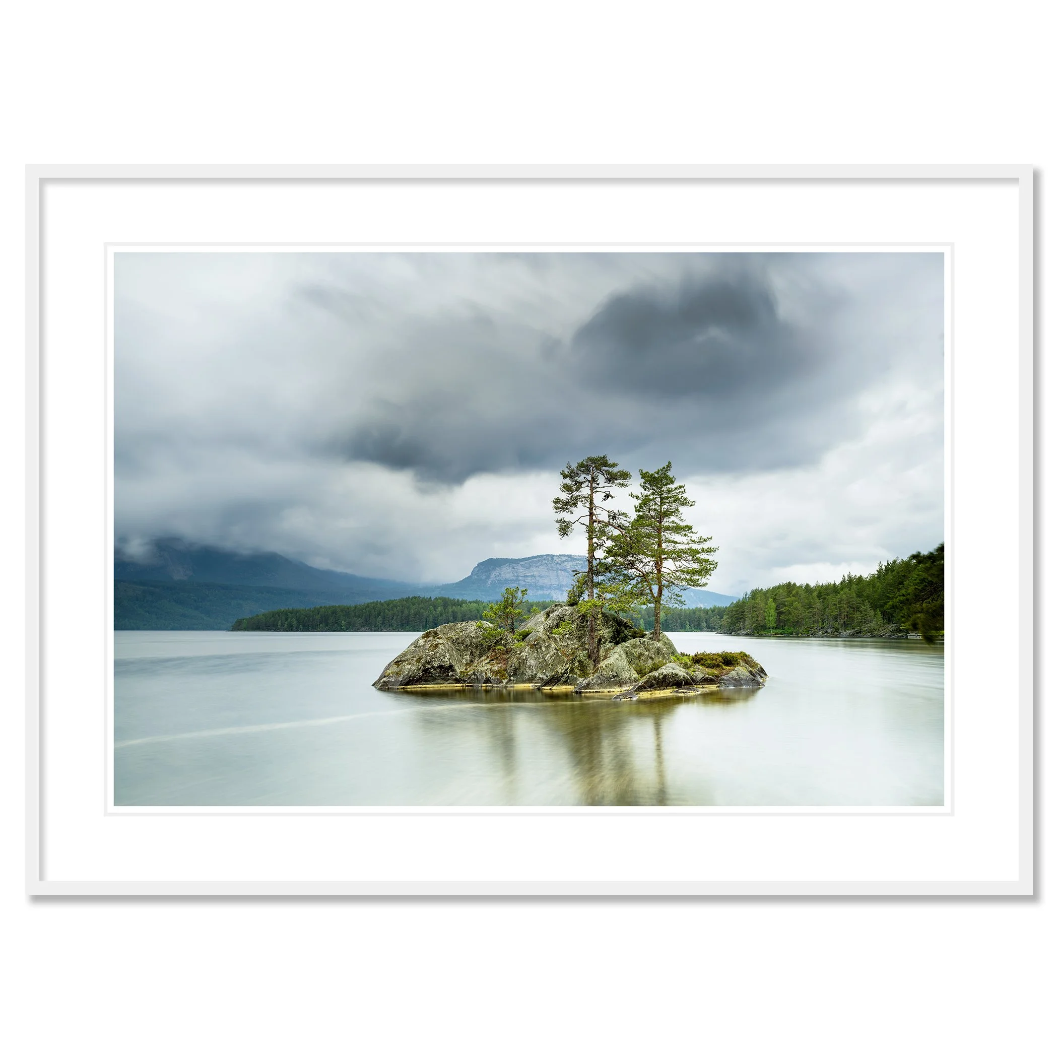 Gerald Berghammer - Color Photography. A small rocky island with tall pine trees surrounded by a calm lake under a cloudy sky and mountains behind. Classic framed white