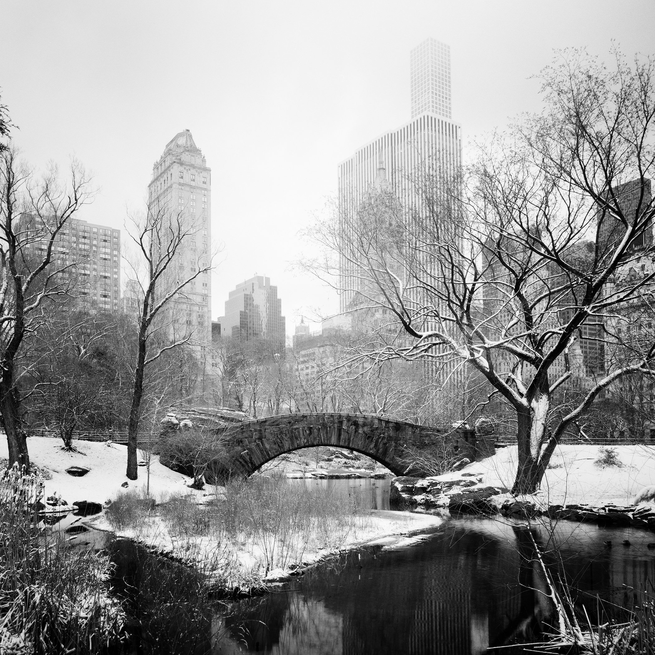 Black and white photograph of a snow-covered bridge in Central Park with Manhattan skyscrapers behind