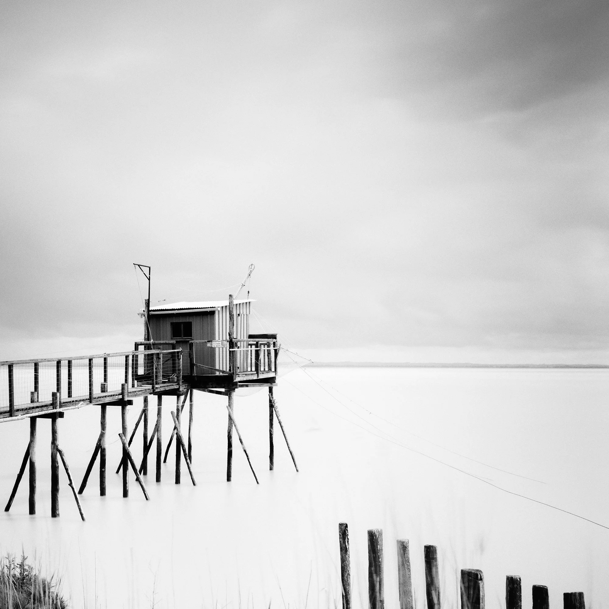 Gerald Berghammer - Black and white seascape photography. Small wooden hut on stilts connected by a long walkway over water with grass and a cloudy sky. Print detail 2