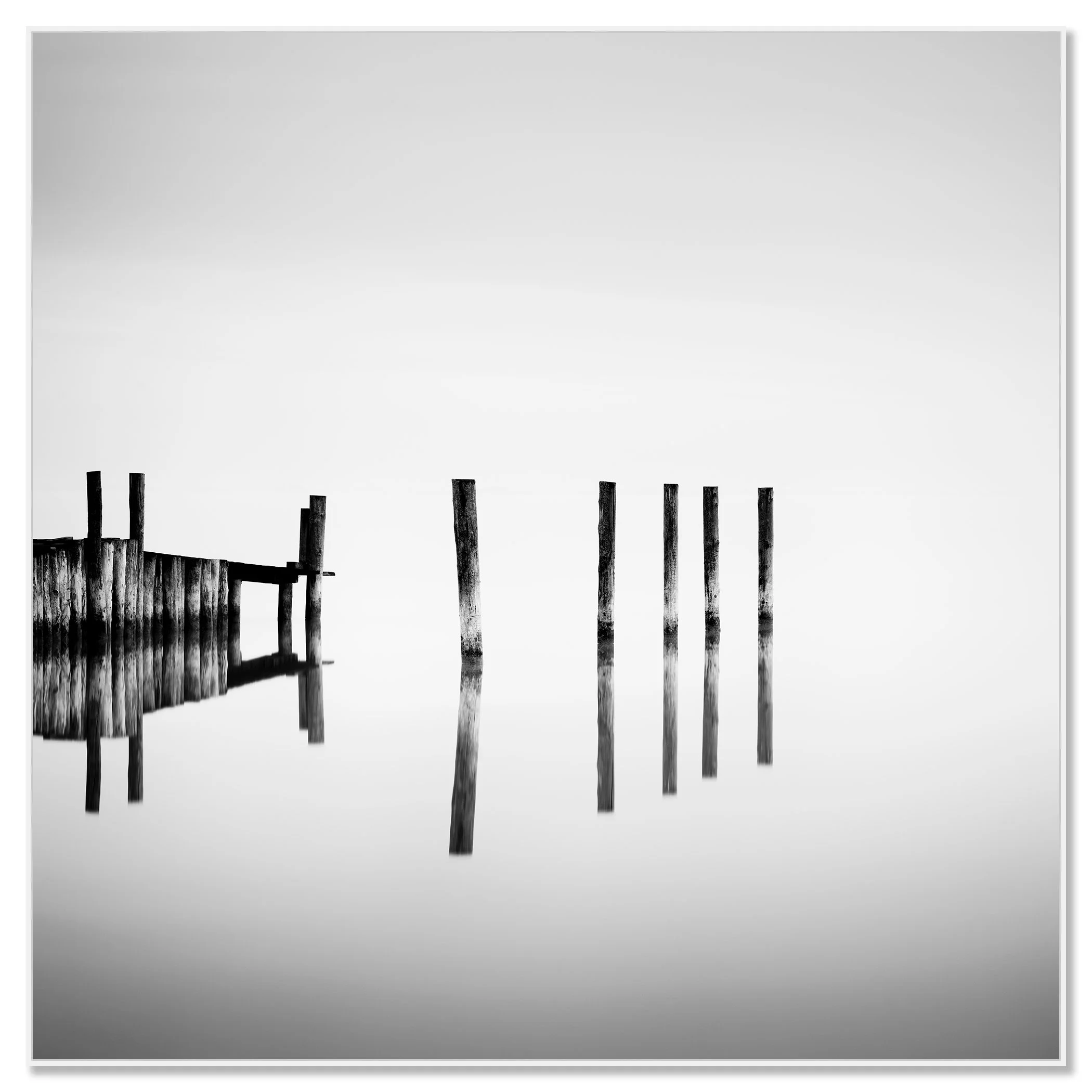 Black-and-white photo of weathered posts and dock reflected in calm lake water beneath an overcast sky – framed ArtBox white