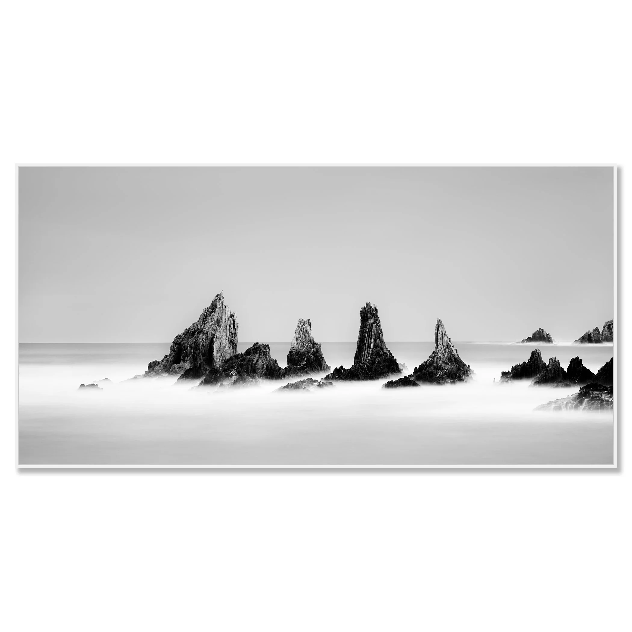 Black-and-white long-exposure photograph of jagged rocks in calm ocean water, misty waves and clear sky – framed ArtBox white