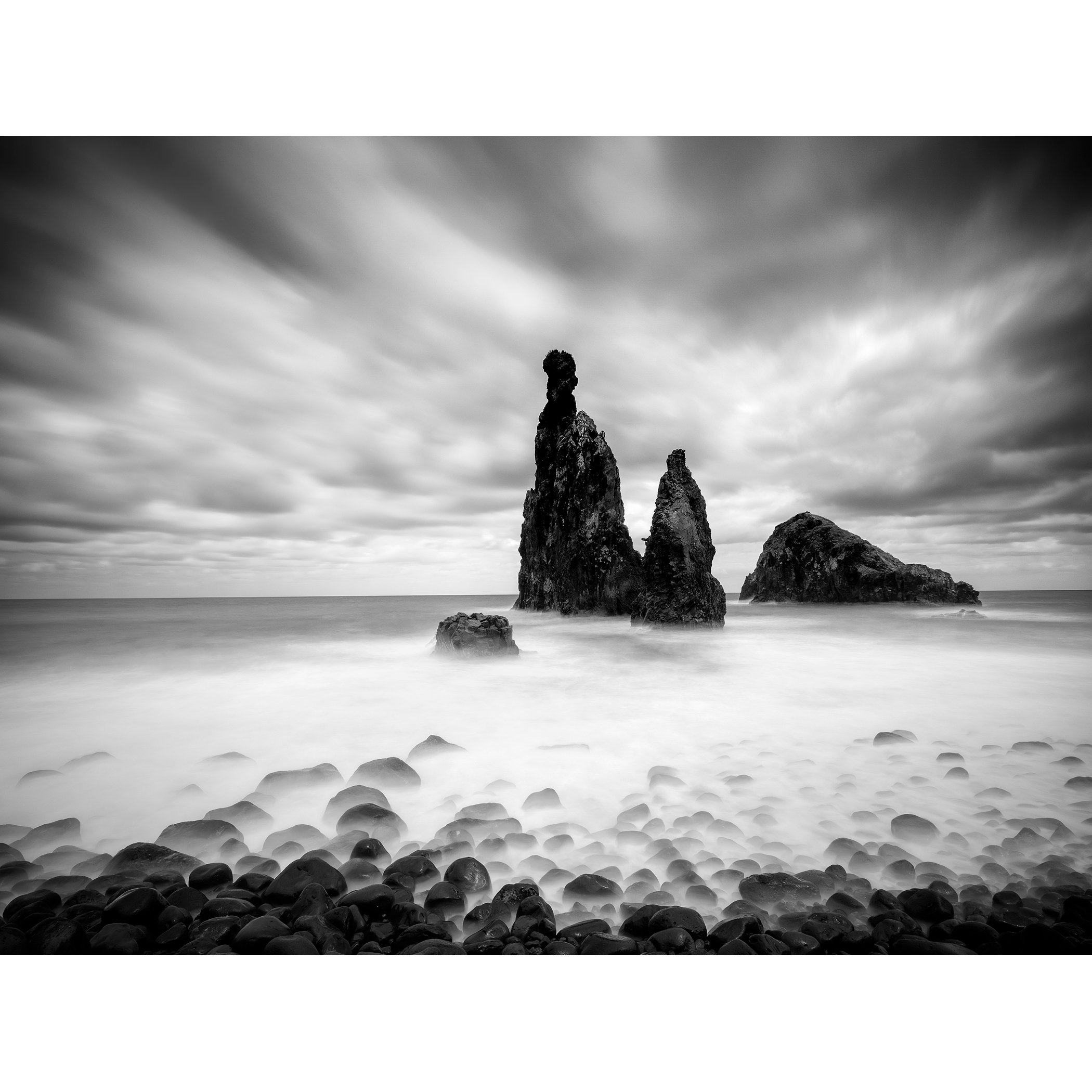 © 2021 Gerald Berghammer - Black and white seascape photography. Rocky seashore with three prominent sea stacks rising from the water, cloudy sky overhead.