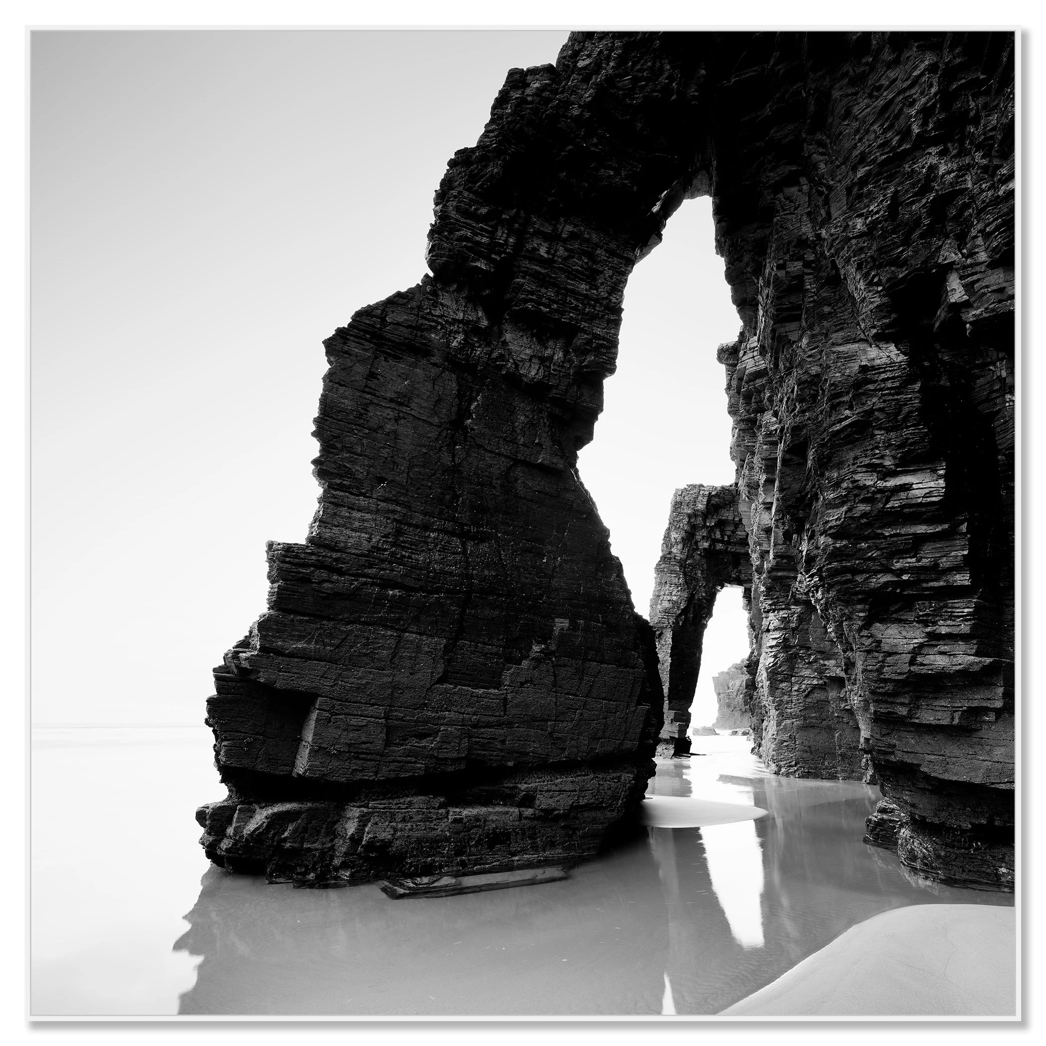 Black and white photo of dramatic sea arches and layered rock cliffs on a quiet sandy beach – framed ArtBox white
