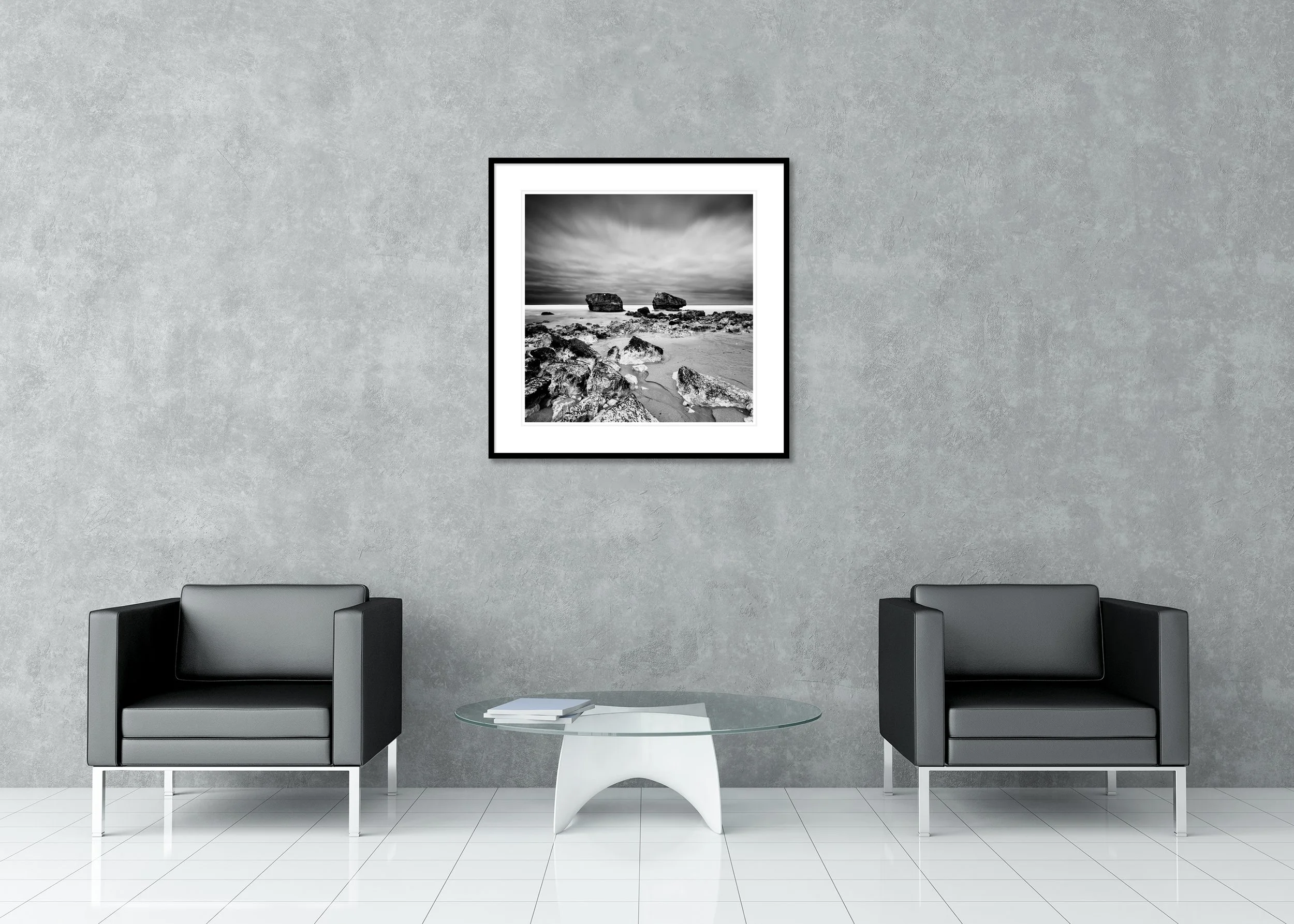 Monochrome seascape archival pigment print with stones on sand, two offshore rocks and a moody sky, presented in a classic frame with a mount.