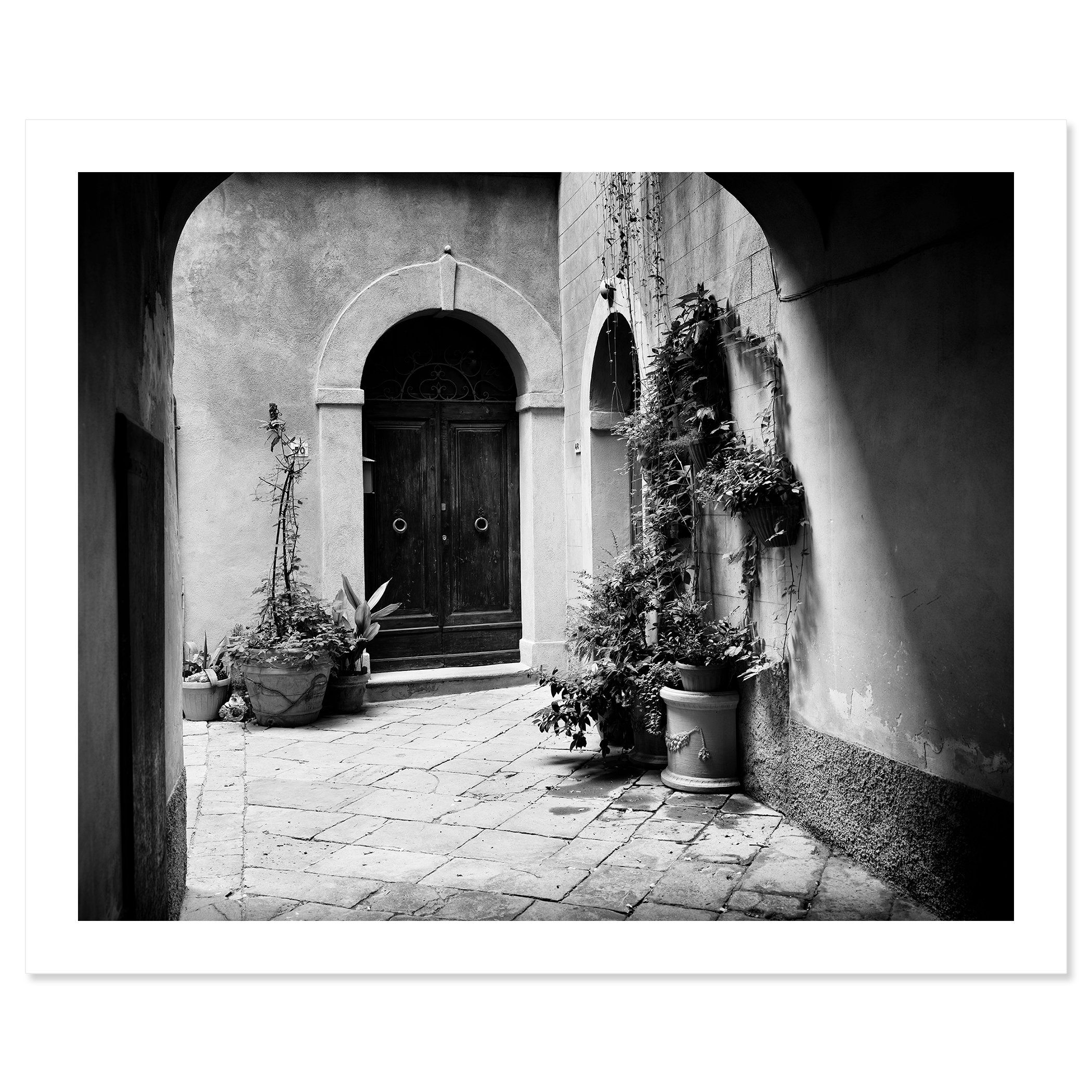 Gerald Berghammer - Black and white cityscape photography. A courtyard with a wooden door, arched doorways, and potted plants on a cobblestone floor. Fine art print only