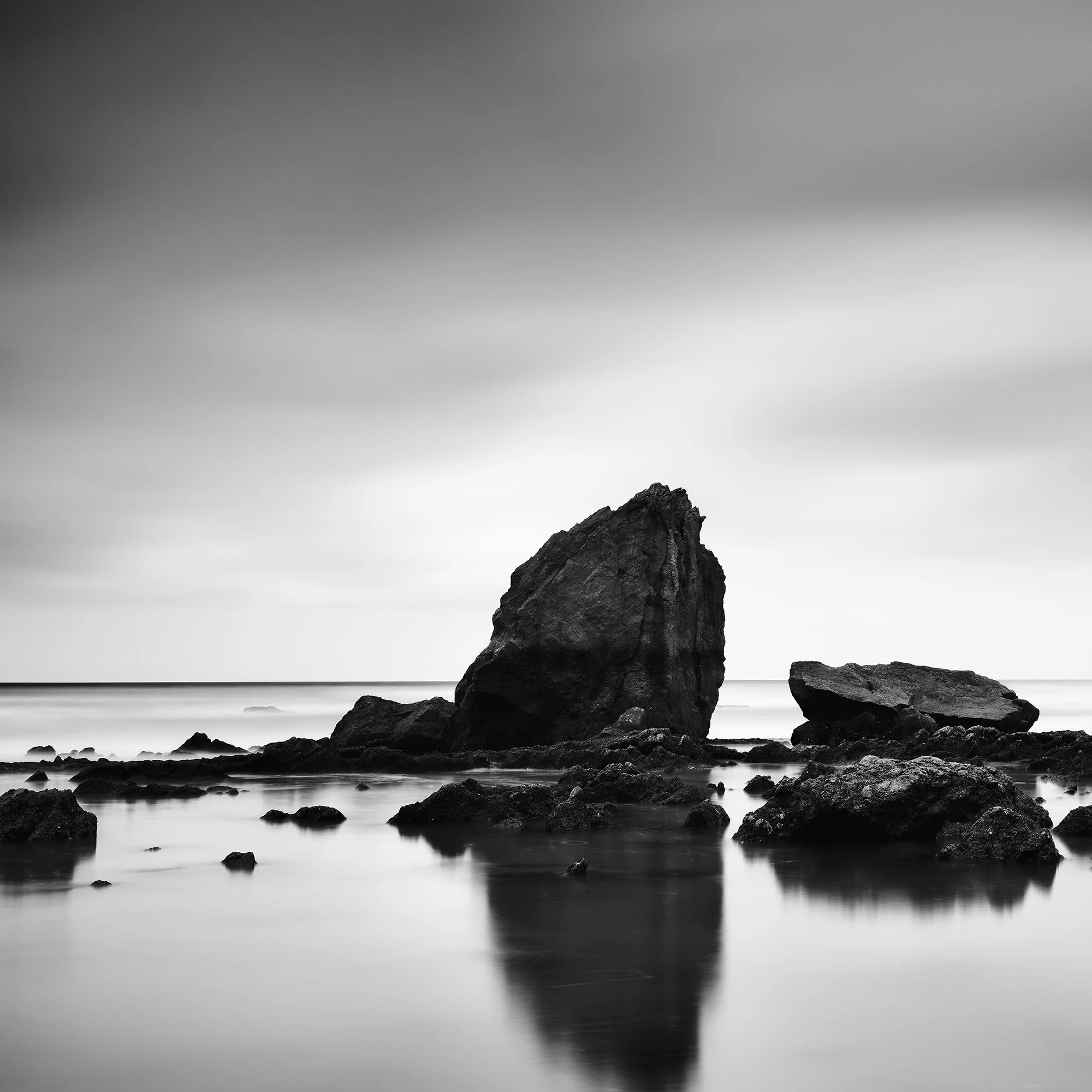 © 2023 Gerald Berghammer - Black and white minimalist beach seascape. A rocky shoreline with a large prominent rock formation in the water. Print detail 1