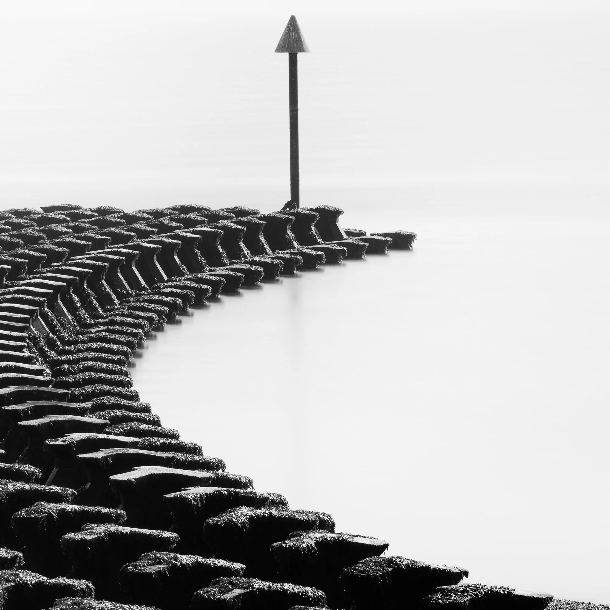Gerald Berghammer - Black and white minimalist photography. A curved stone jetty extending into a calm body of water, with a tall navigation marker at the end. Print detail 3