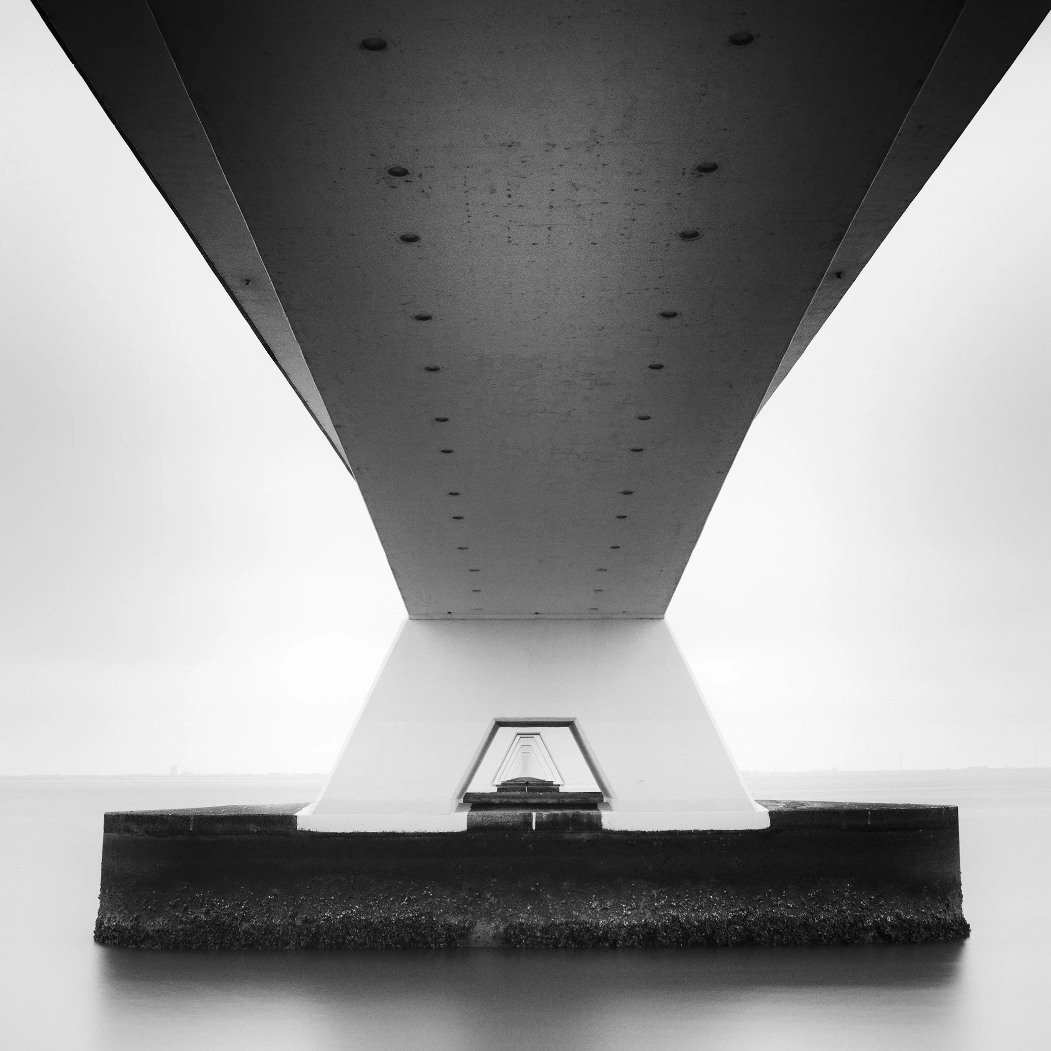 Gerald Berghammer - Black and white cityscape photo. Underneath view of a bridge with large support pillars, showing the underside and the water below.