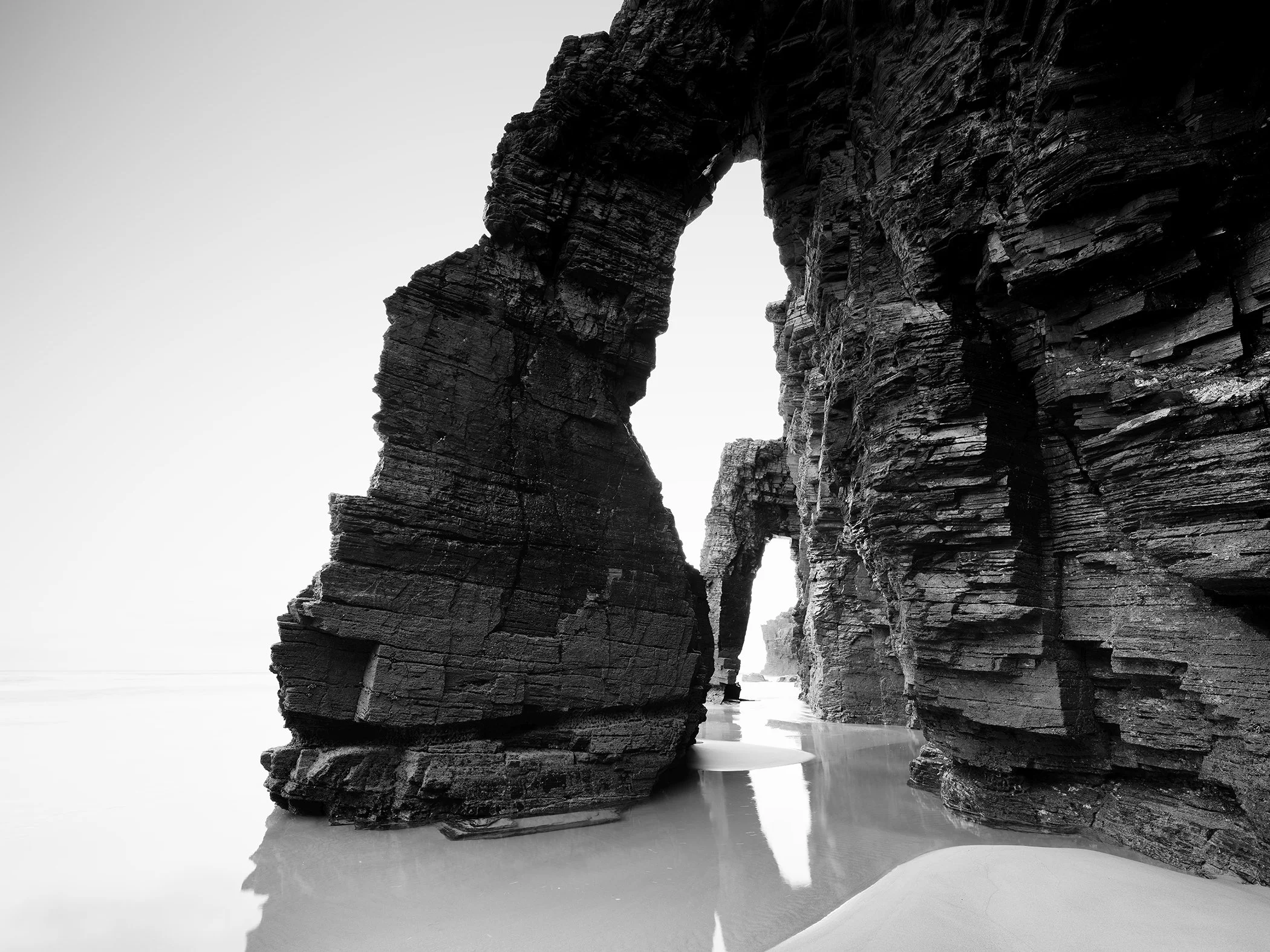 Black and white seascape photography of large natural rock arches on a beach, with calm water reflecting the formations