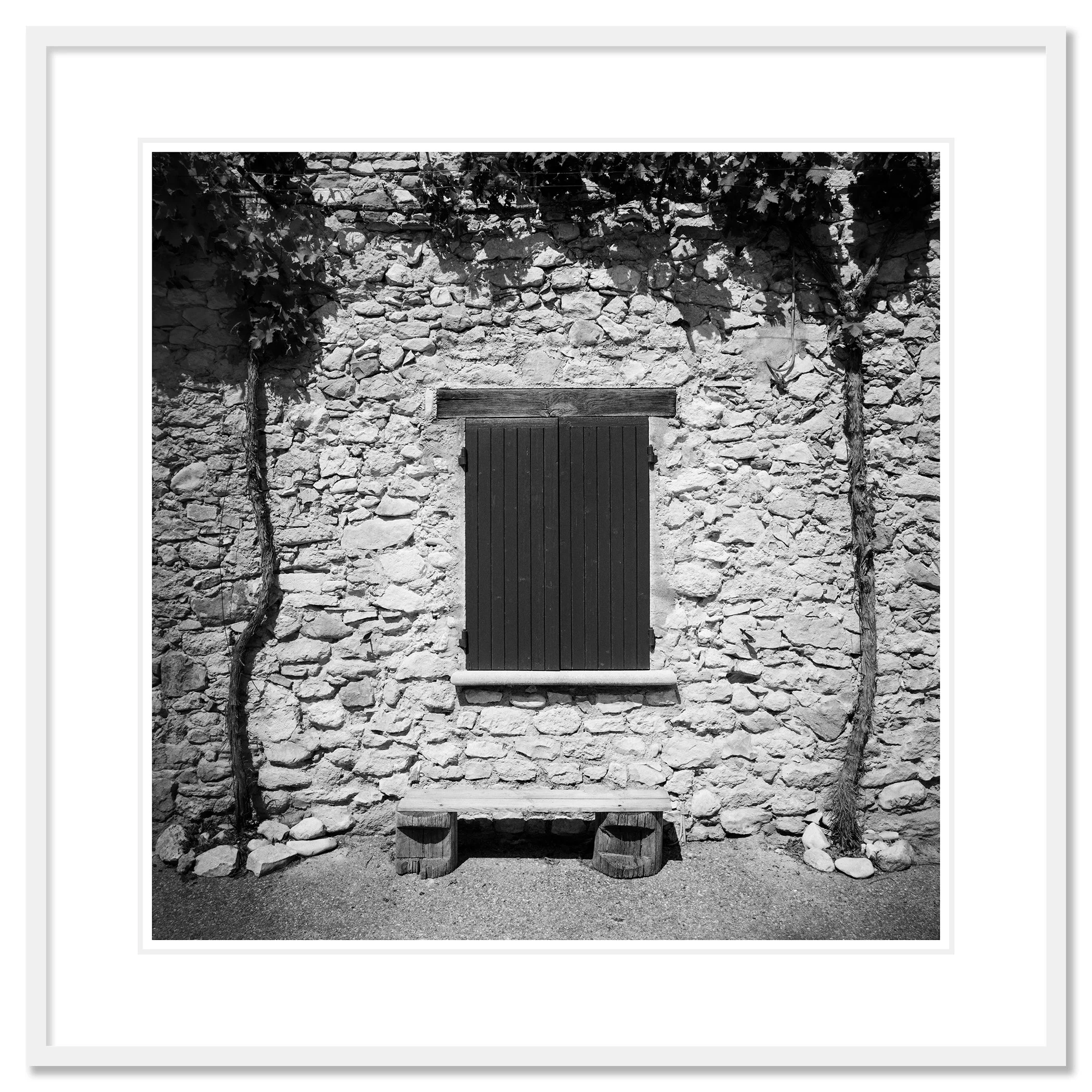Gerald Berghammer - Black & white photography. Stone wall with a closed wooden window shutter, flanked by climbing vines on both sides, and a small bench. Classic framed white