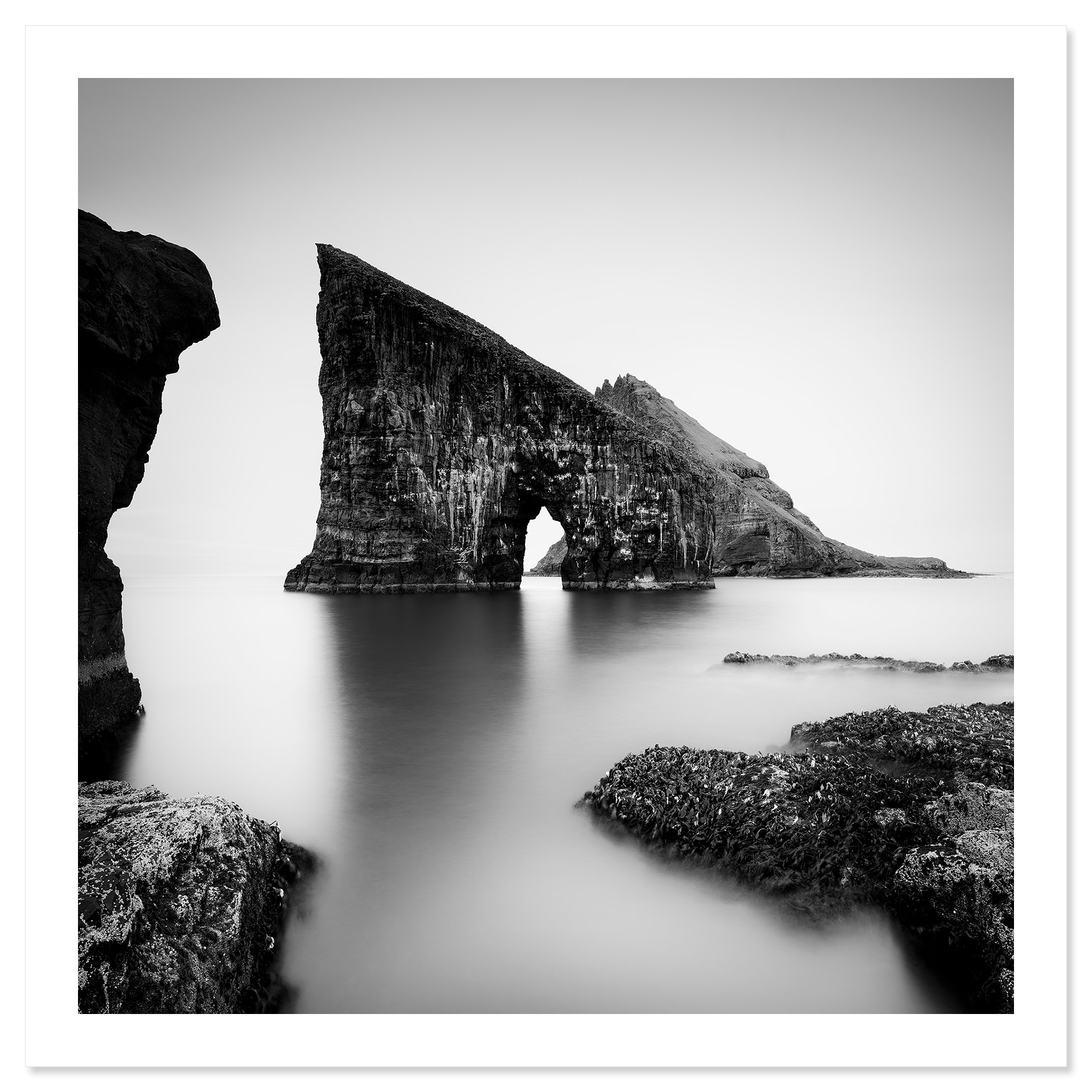 Black and white coastal landscape with a striking sea stack arch and soft long-exposure water – print only