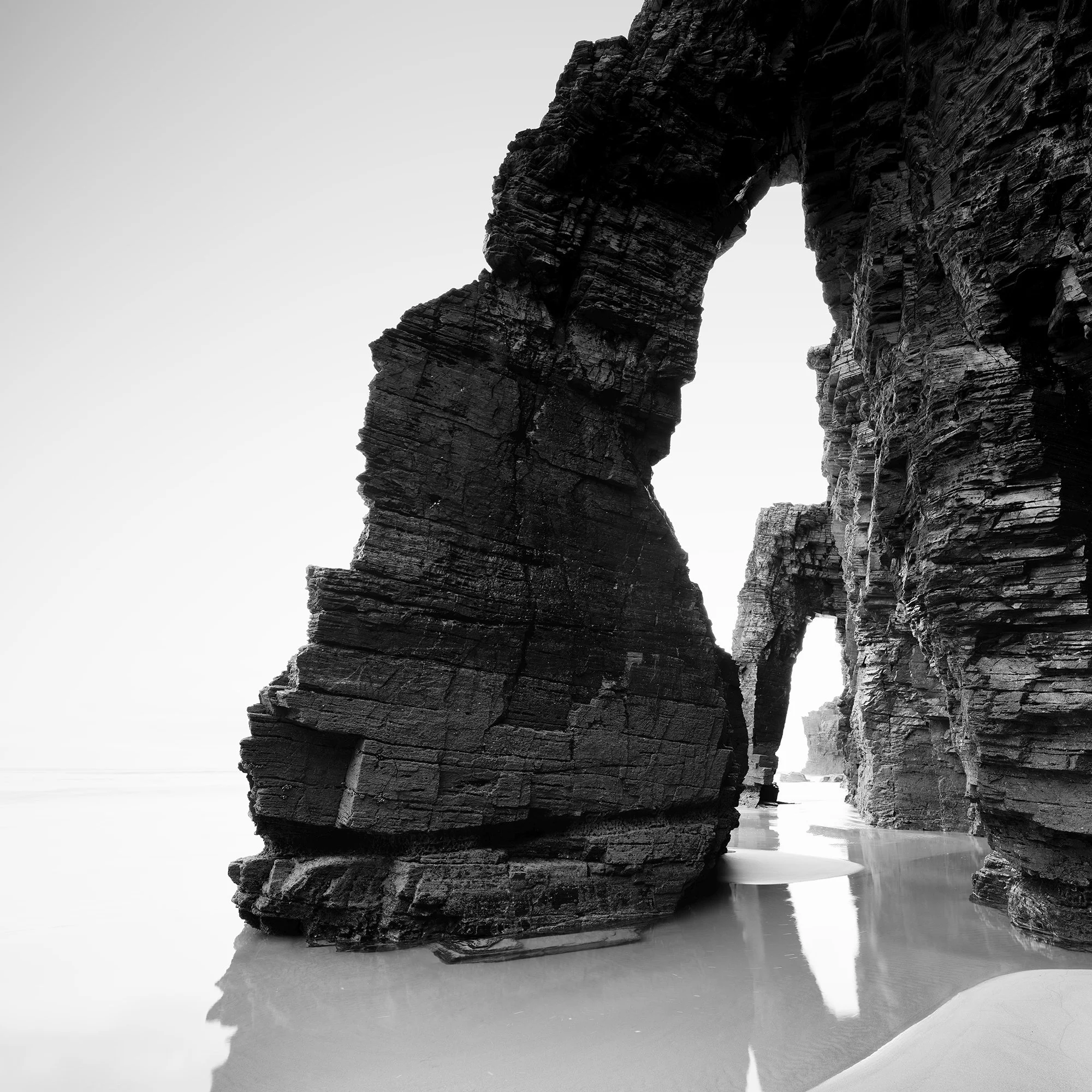 Gerald Berghammer - Black and white beach seascape photography. Large natural rock arch formations on a beach with water reflecting the rocks. Print detail 1