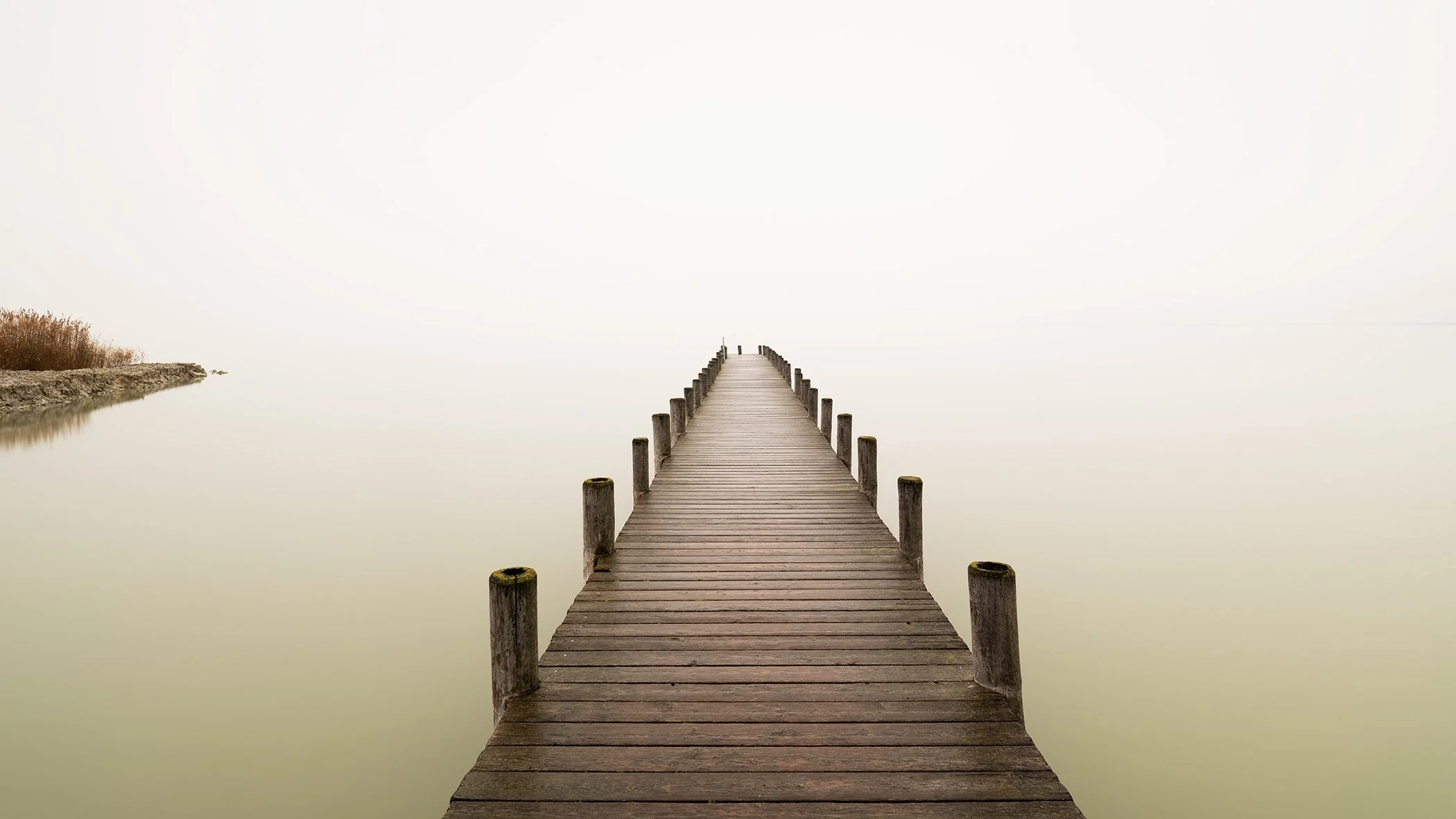 Minimalist photograph of a wooden pier extending into a calm, mist-covered lake, photographed by Gerald Berghammer.
