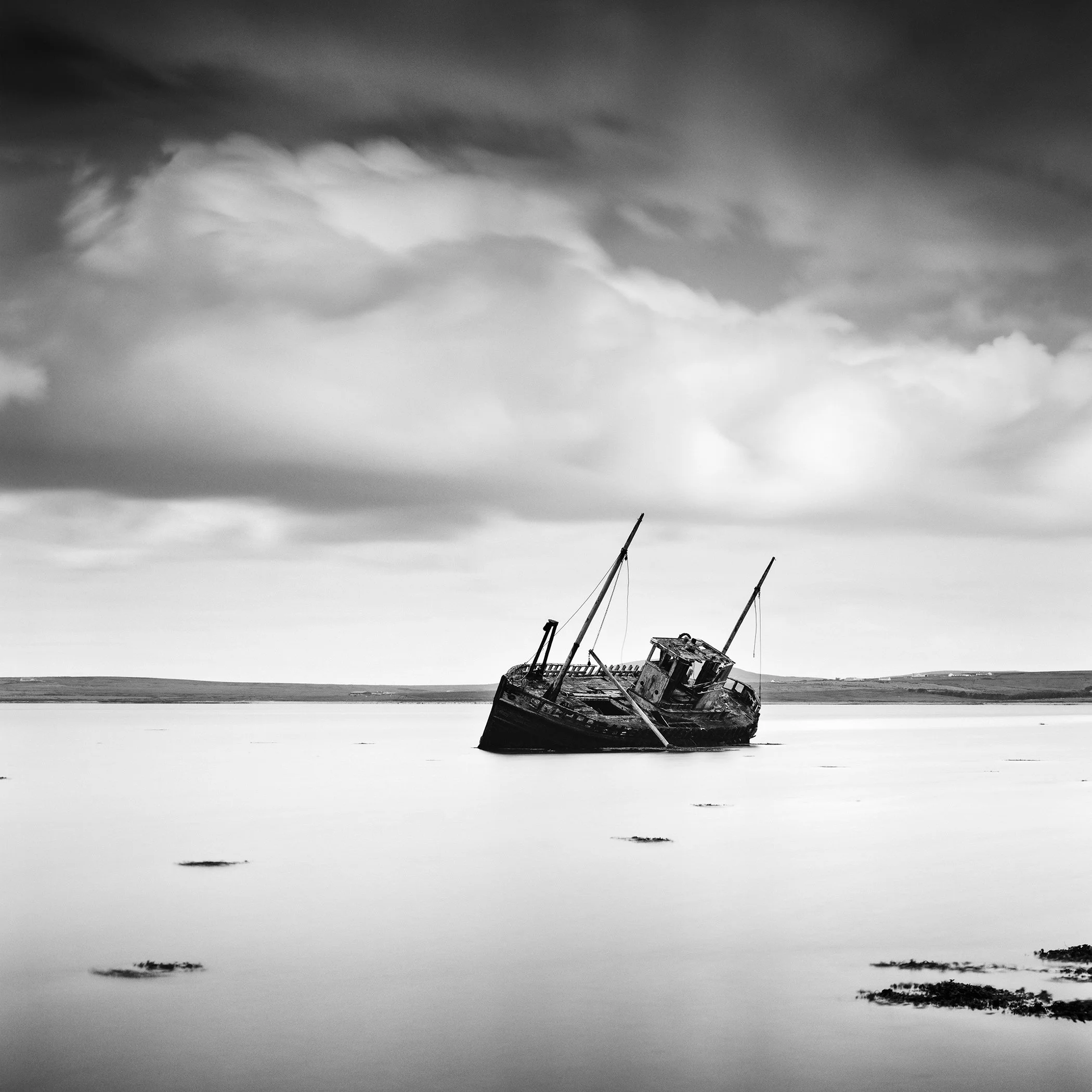 Black-and-white photo of an abandoned boat resting on calm water in Ireland beneath dramatic clouds