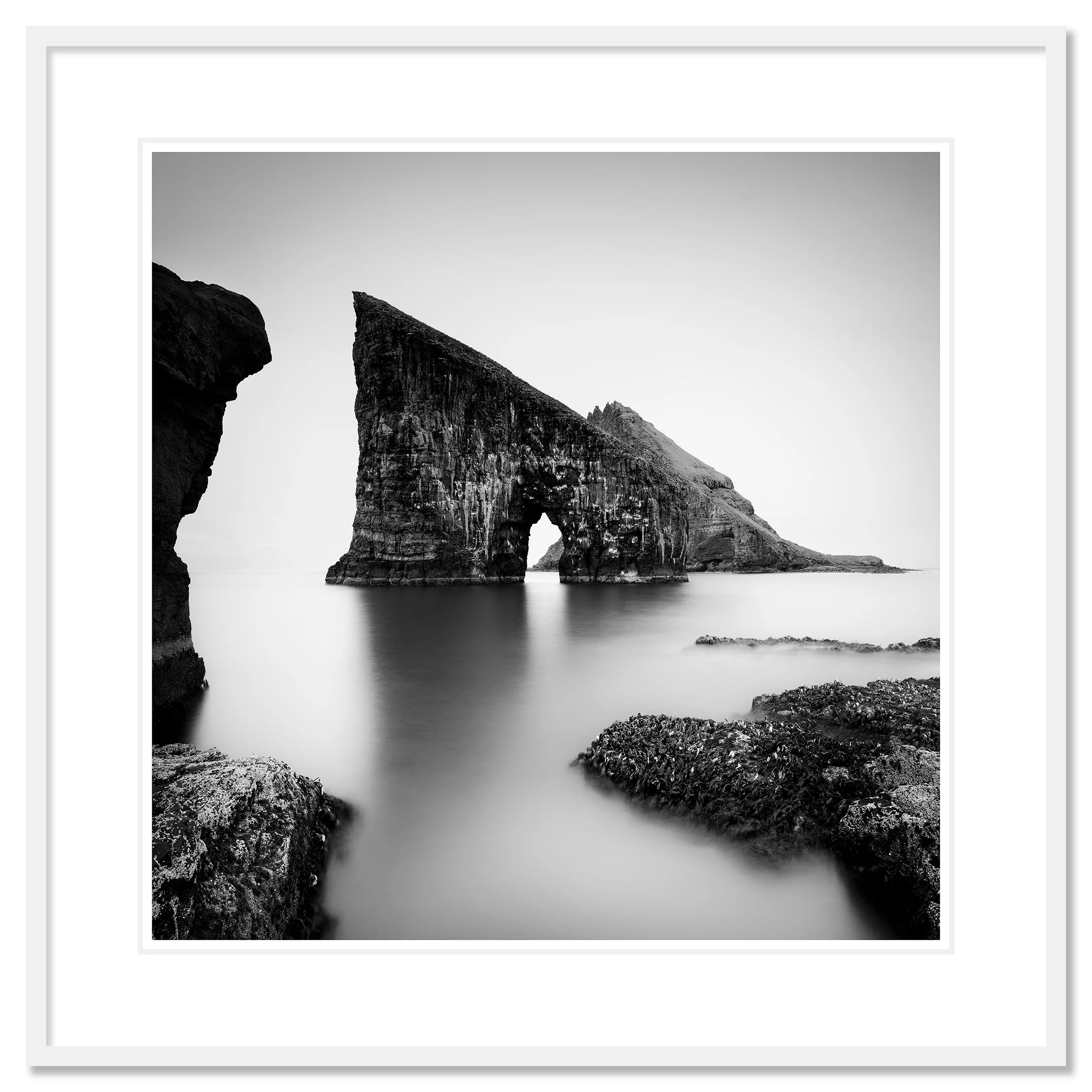 Black and white coastal landscape with a striking sea stack arch and soft long-exposure water – classic framed white