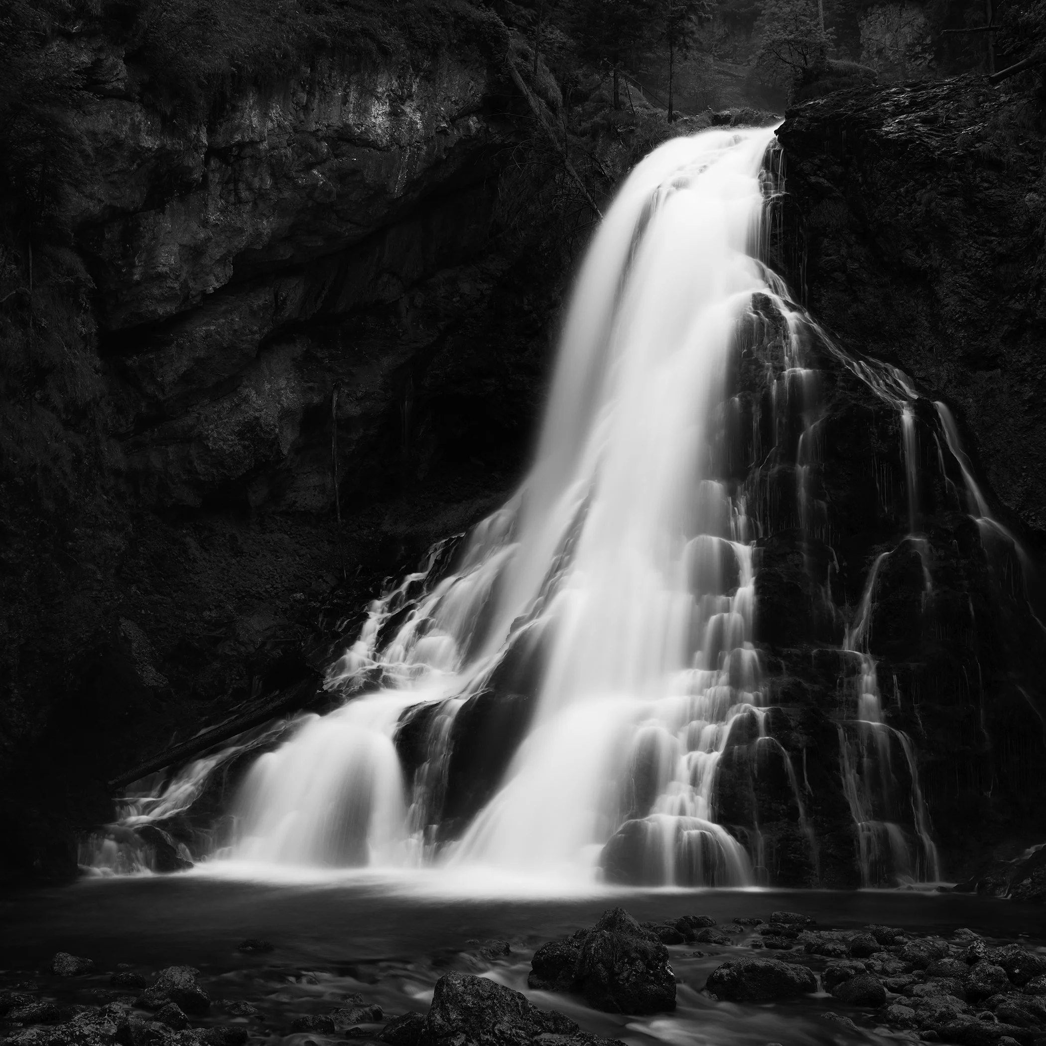 Black-and-white long-exposure photo of Golling Waterfall cascading over dark rocks into a calm pool, surrounded by rugged natural scenery.